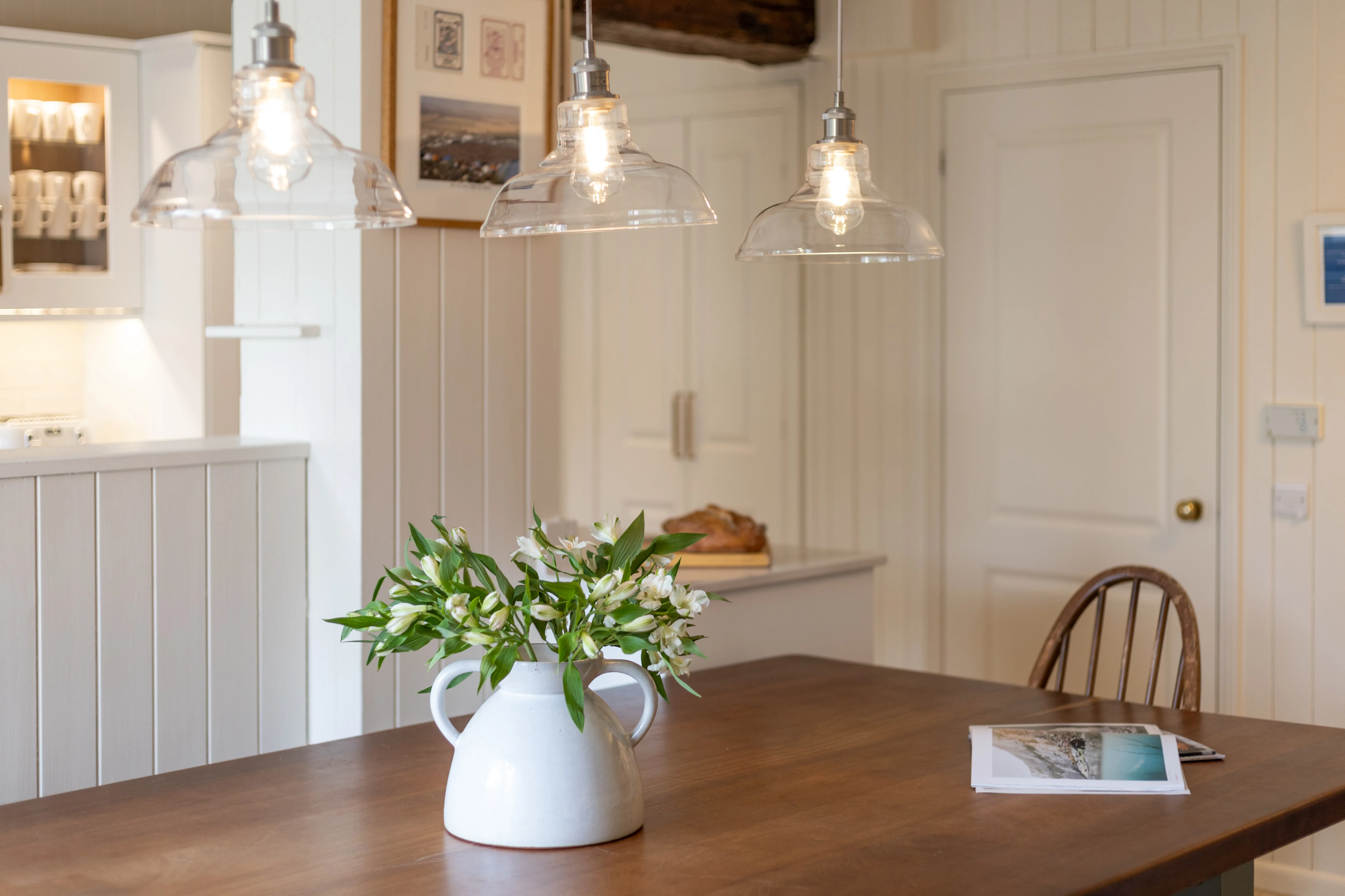 Wooden dining table with a white ceramic jug of flowers beneath glass pendant lights in a bright kitchen.