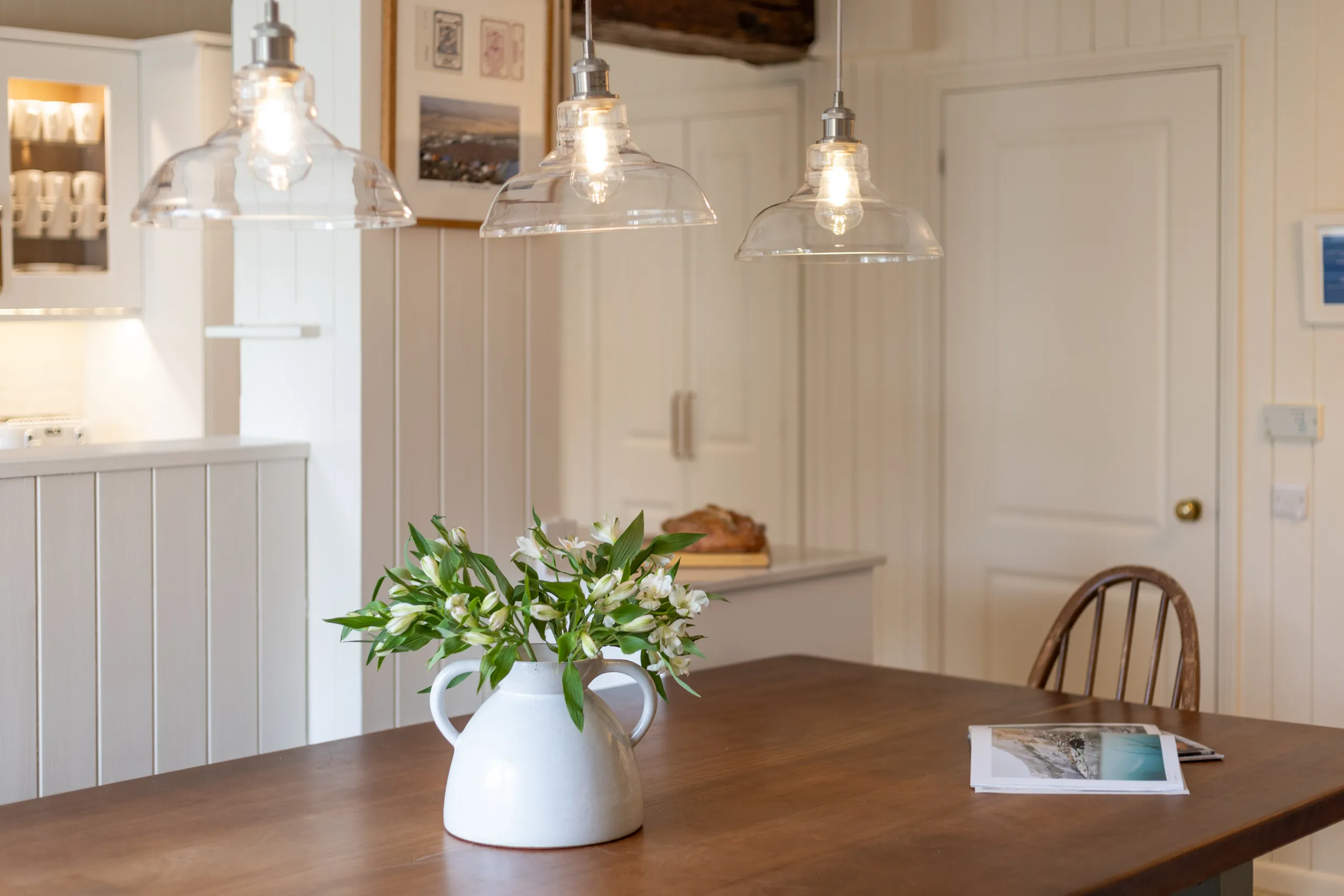 Wooden dining table with a white ceramic jug of flowers beneath glass pendant lights in a bright kitchen.