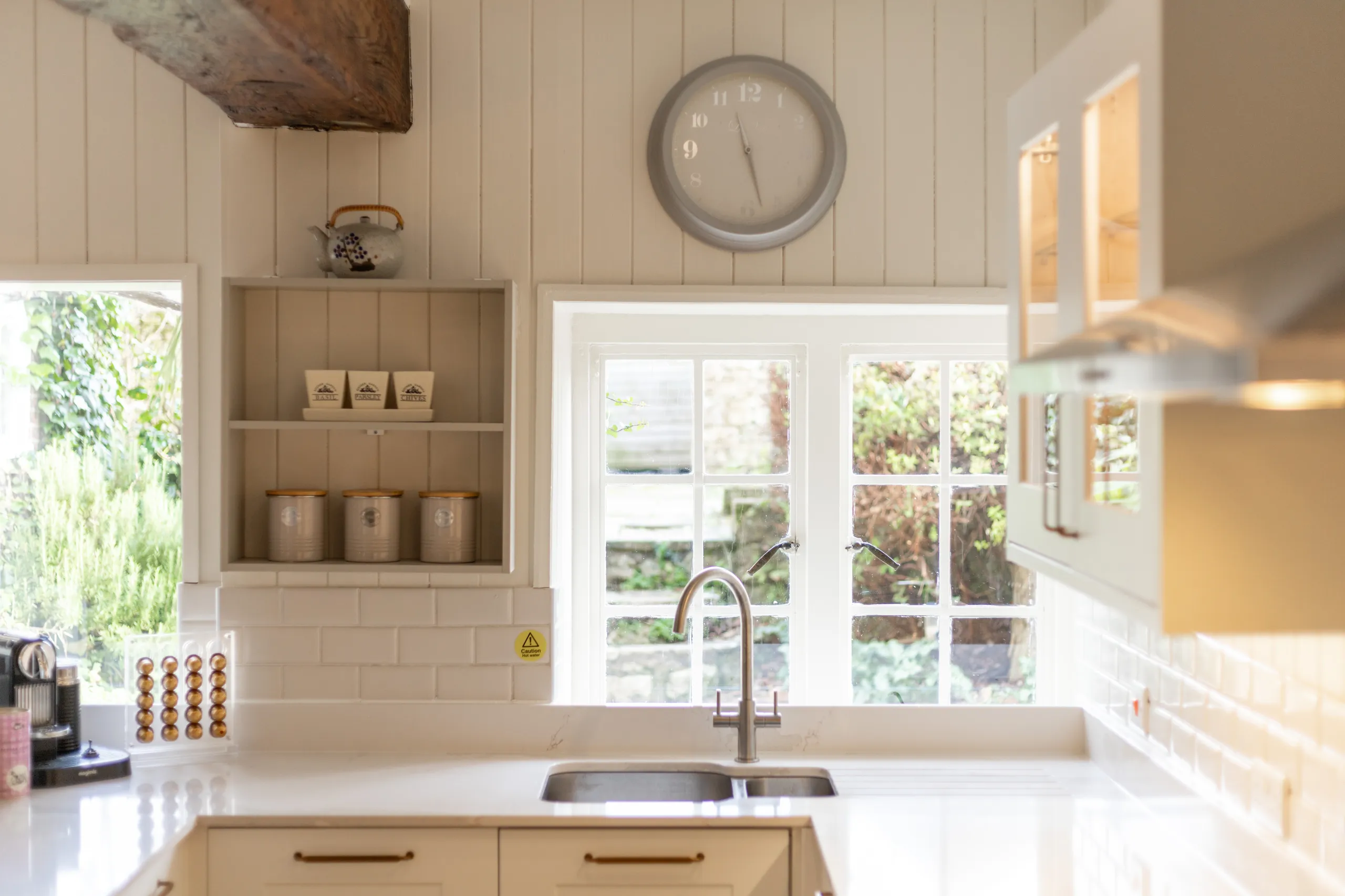 Bright country kitchen sink beneath a window with open shelving, canisters and a wall clock