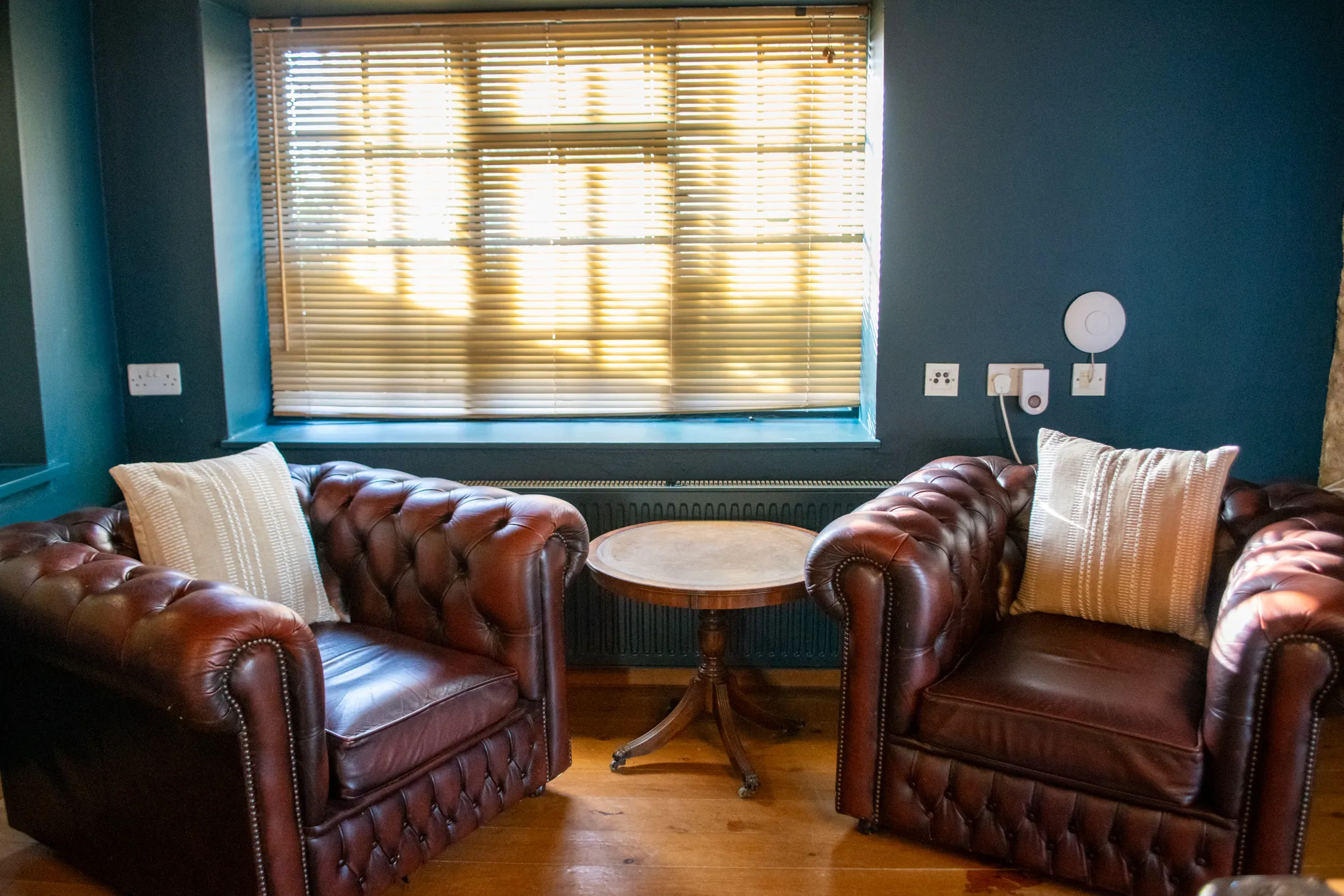 Two brown leather armchairs with cream cushions and a small round table by a window with wooden blinds