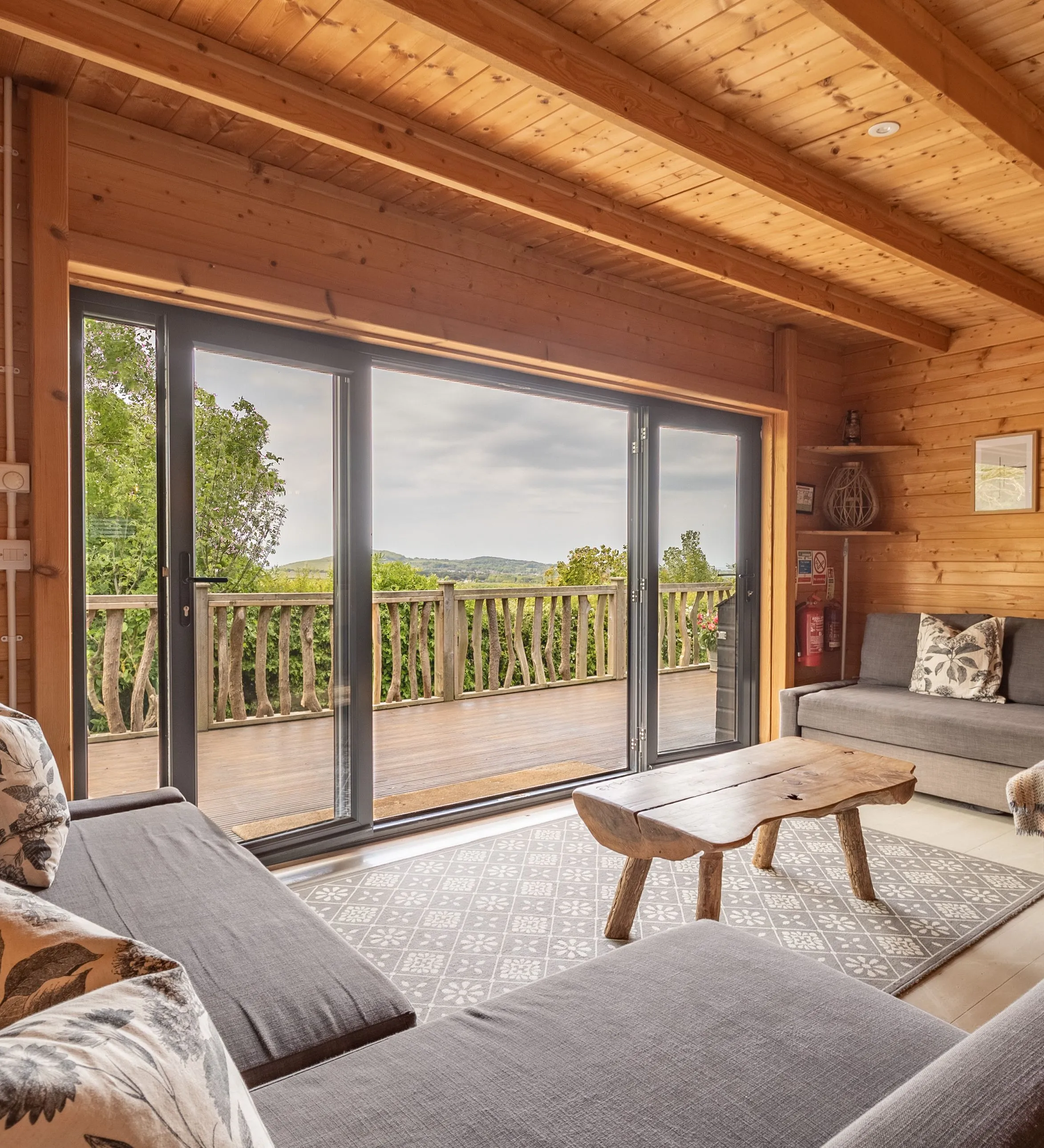 Cosy wooden cabin lounge with grey sofas and sliding doors to a deck overlooking countryside