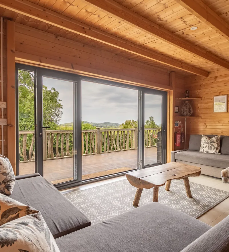 Cosy wooden cabin lounge with grey sofas and sliding doors to a deck overlooking countryside