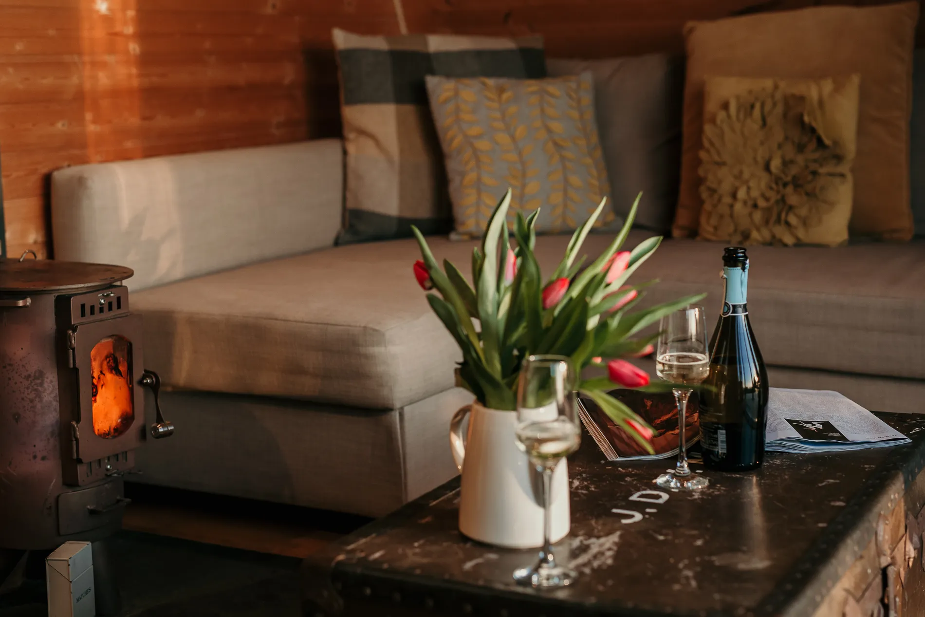 Cosy cabin living area with a wood-burning stove, sofa cushions, and a table with tulips and wine glasses