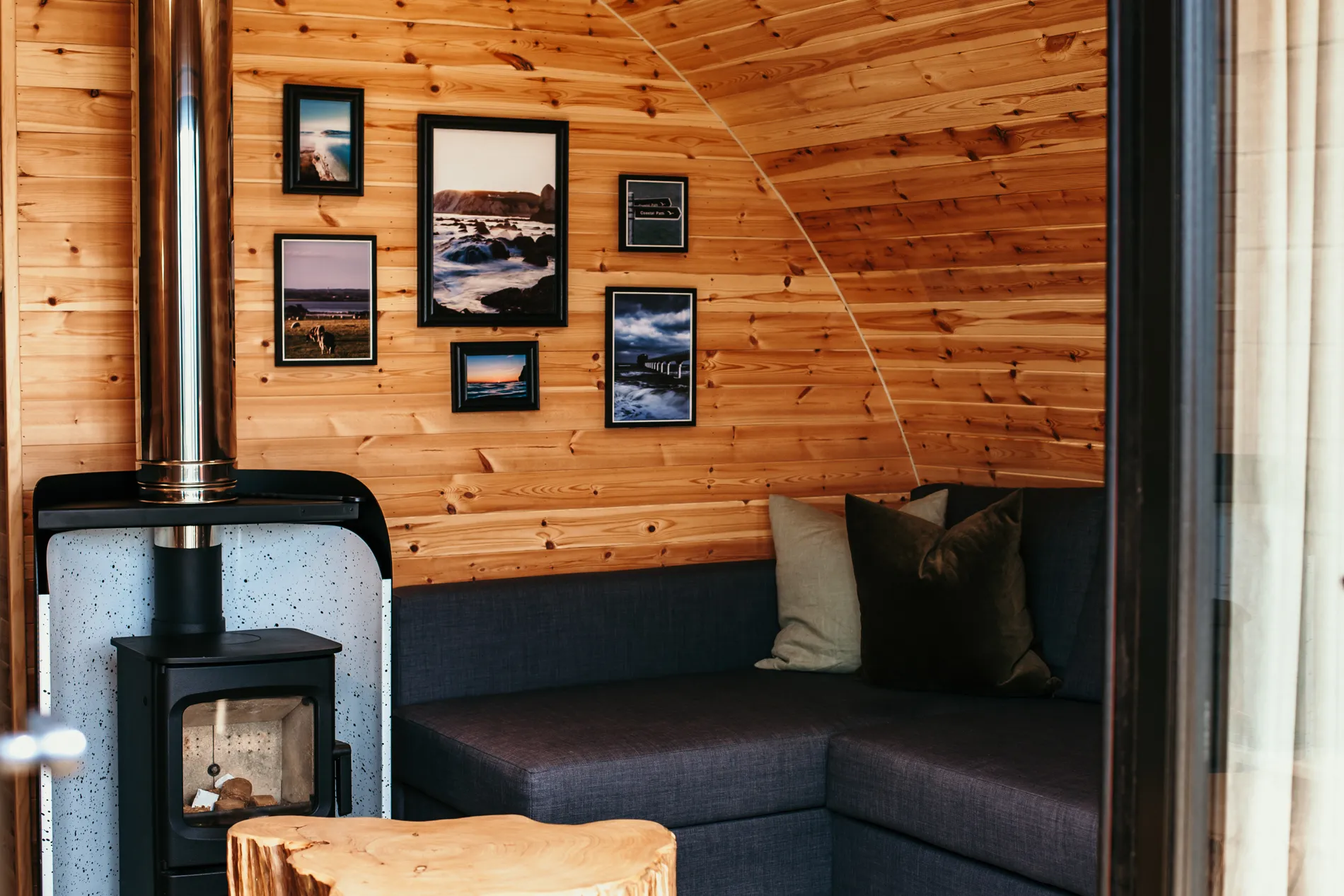 Cosy wooden cabin corner with a log burner, framed photos on the wall and a dark grey sofa with cushions