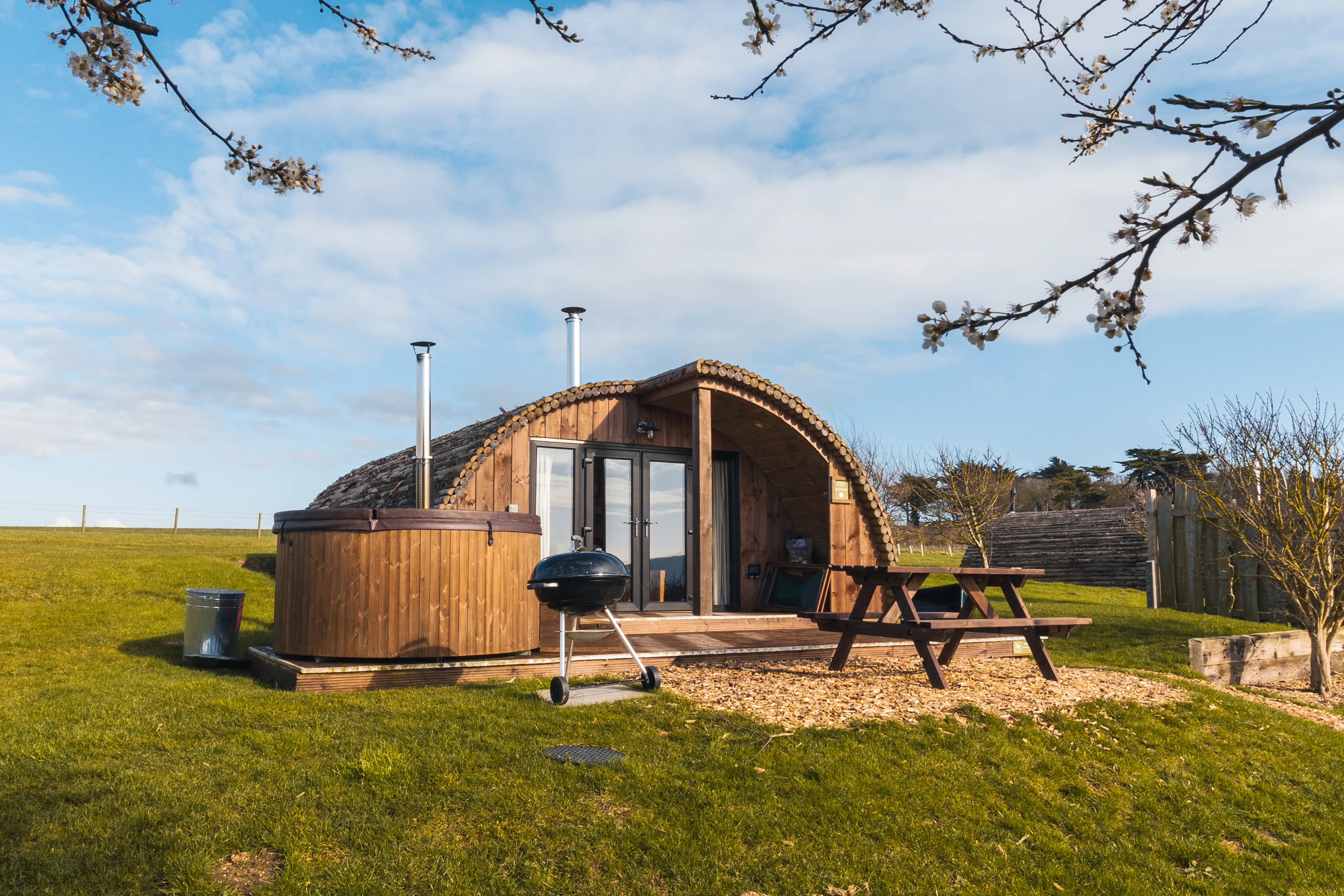 Wooden glamping pod with hot tub and picnic table on a grassy field under a blue sky