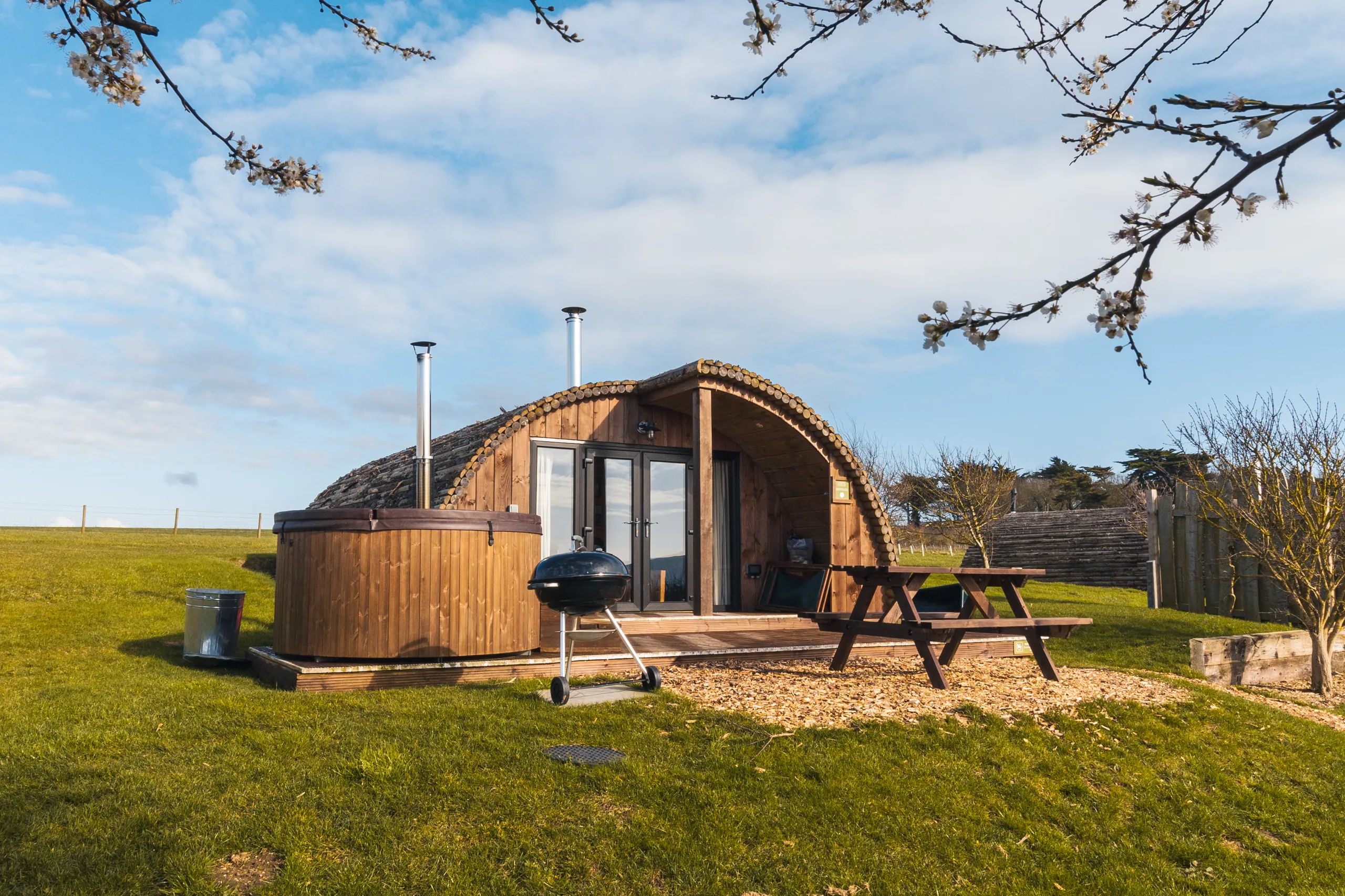 Wooden glamping pod with hot tub and picnic table on a grassy field under a blue sky