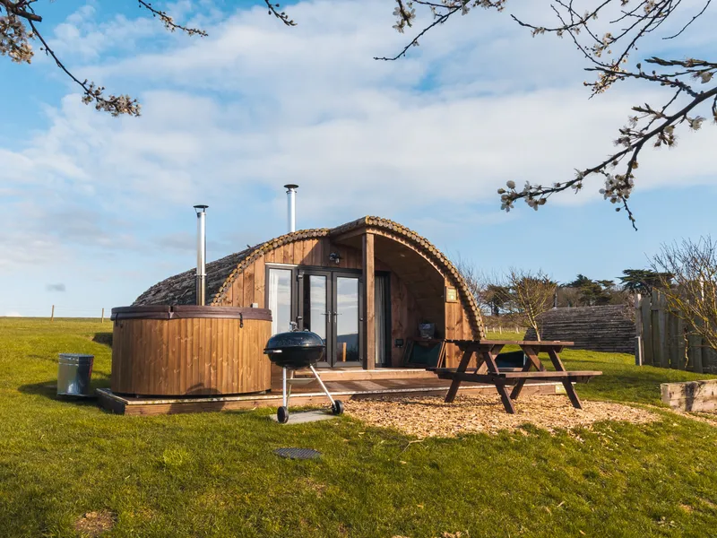 Wooden glamping pod with hot tub and picnic table on a grassy field under a blue sky
