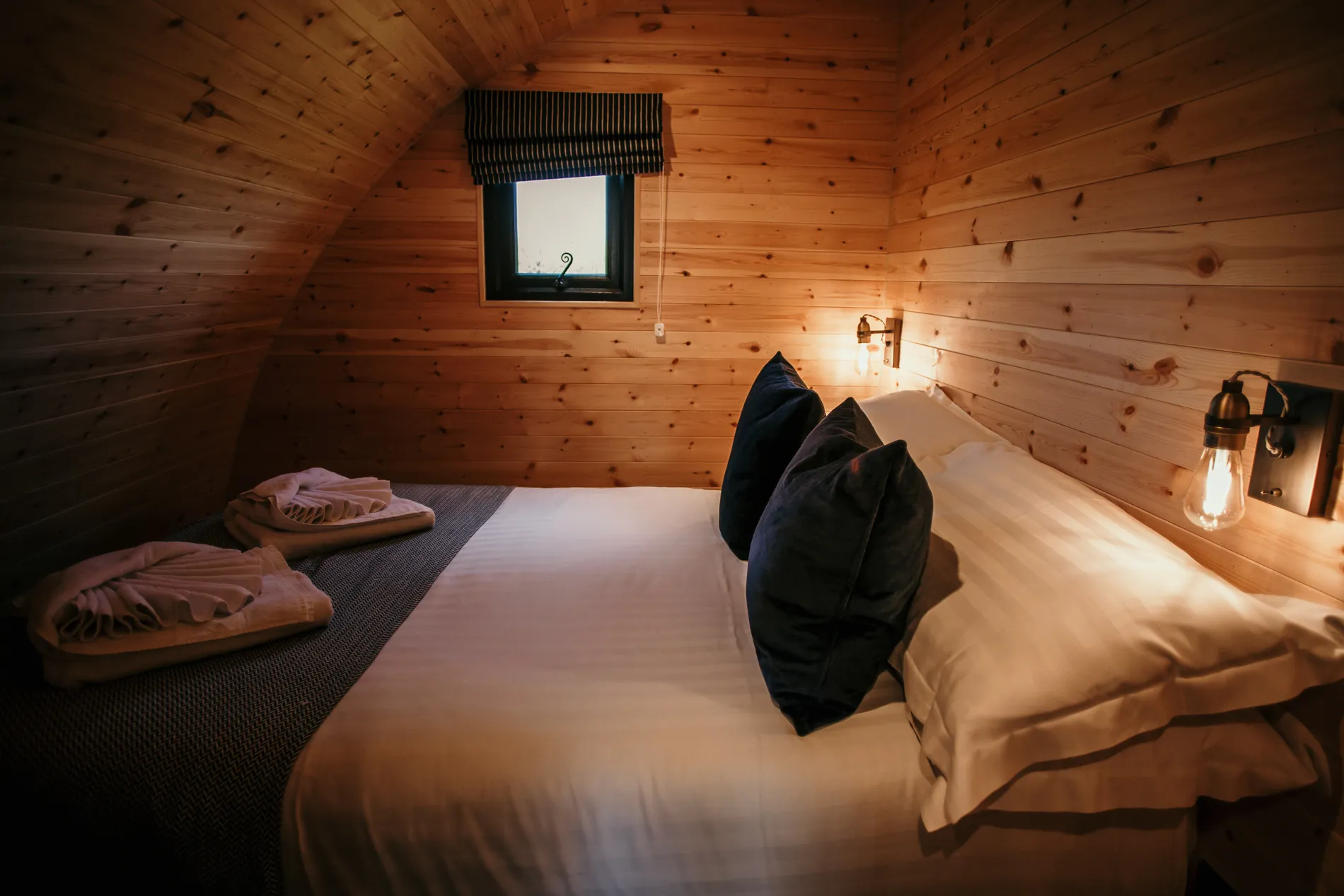 Cosy wooden cabin bedroom with double bed, dark cushions, folded towels and warm wall lights