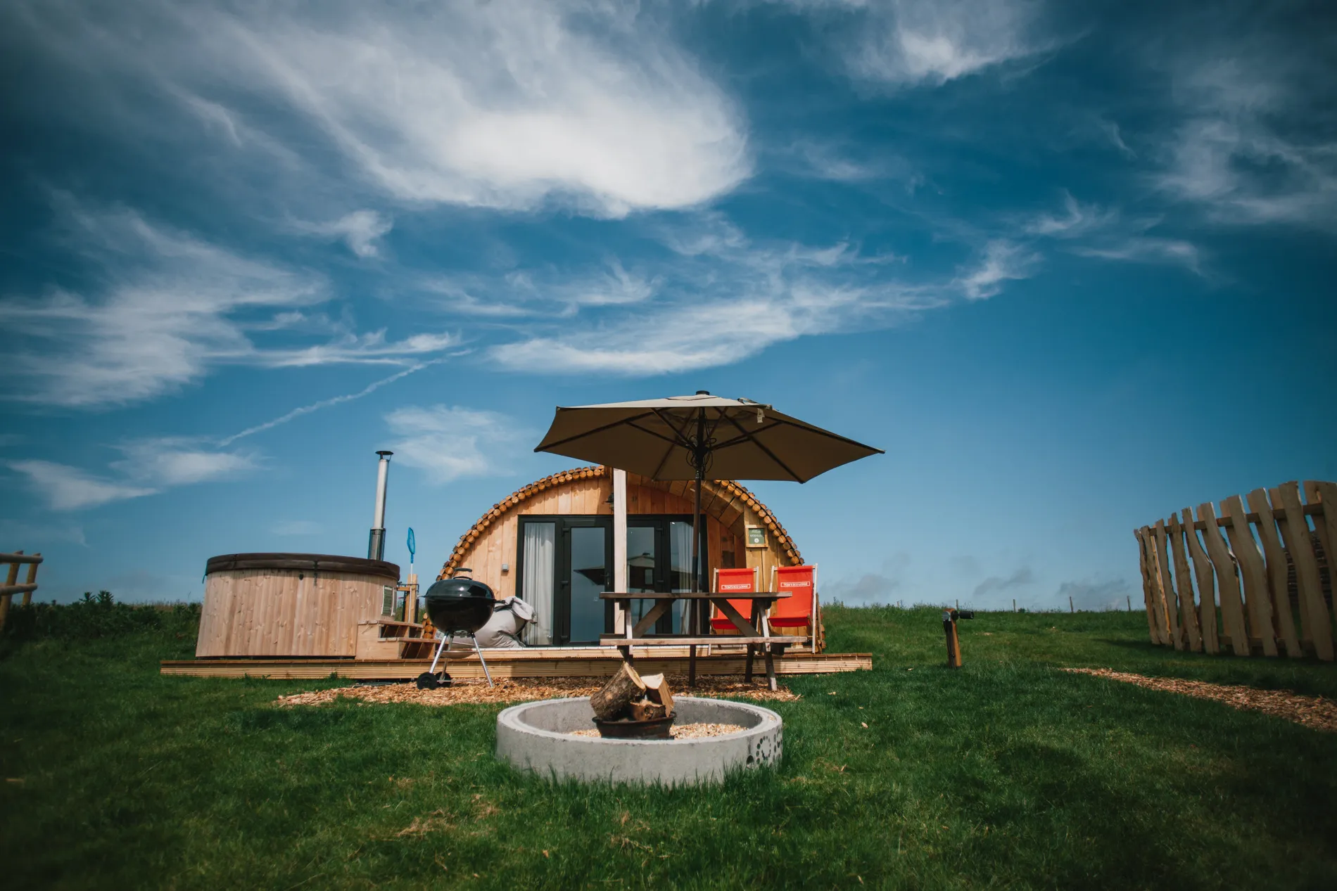 Wooden glamping pod with hot tub and picnic table under a large parasol on a grassy field