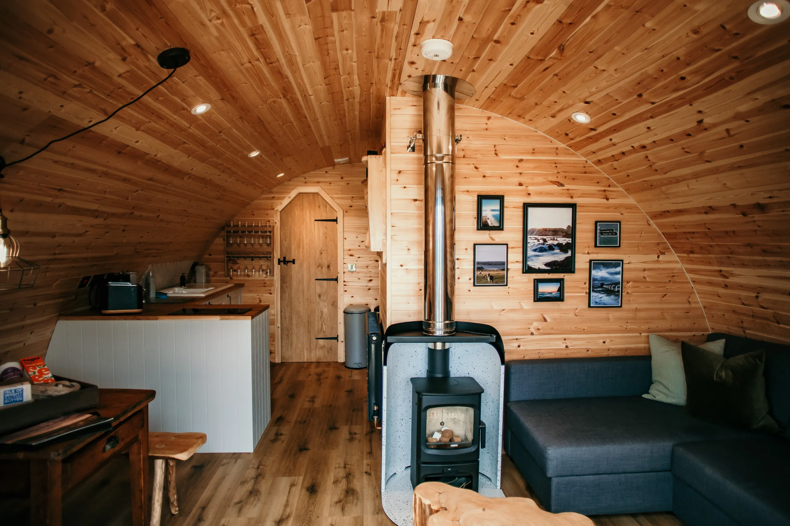 Cosy wooden cabin interior with a wood-burning stove, kitchenette and grey sofa