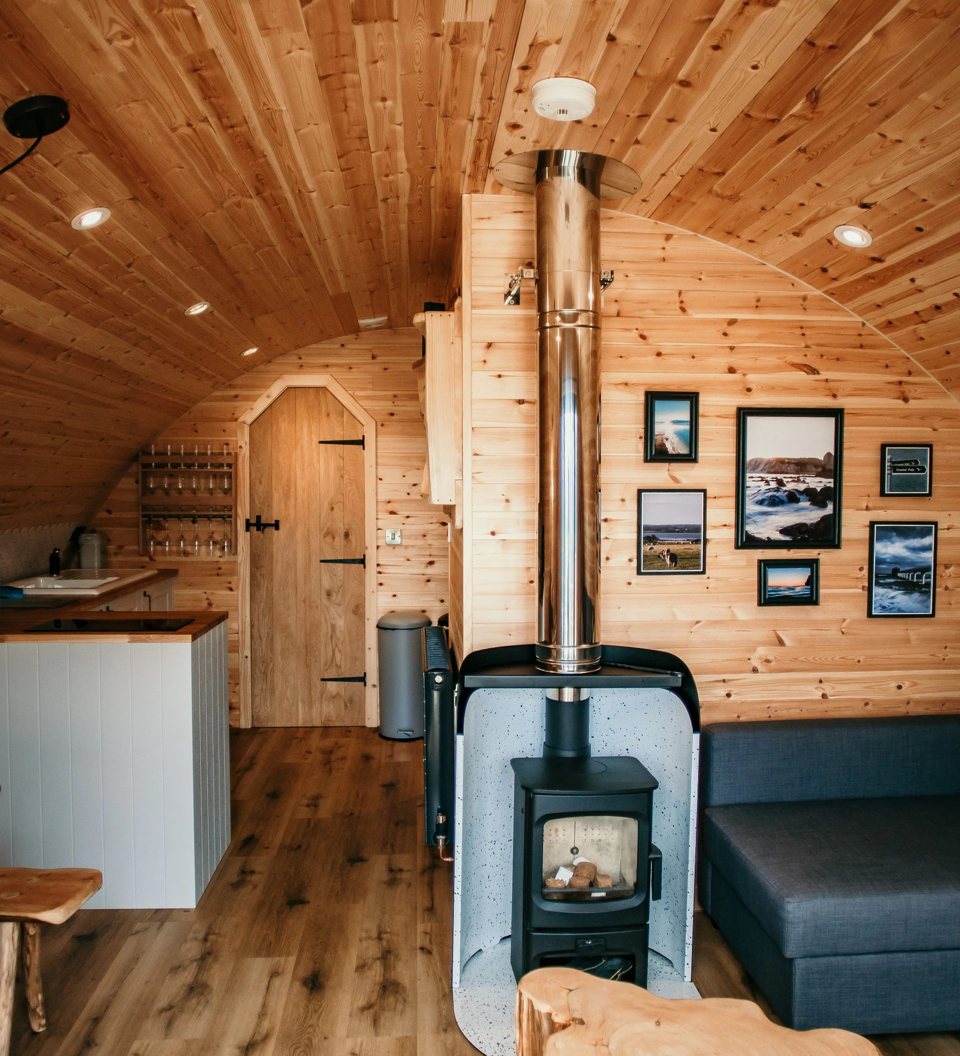 Cosy wooden cabin interior with a wood-burning stove, kitchenette and grey sofa