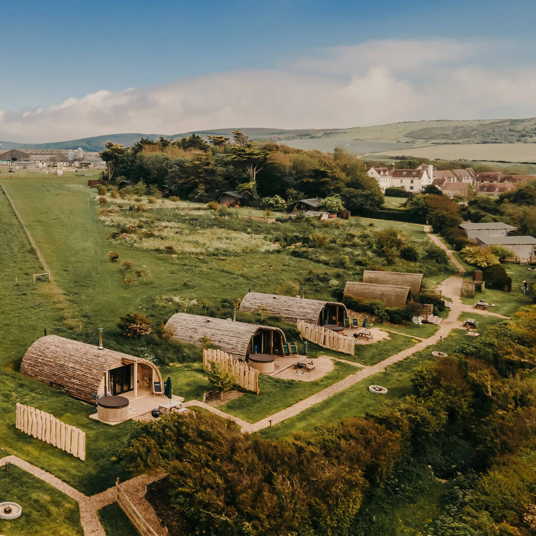 Aerial view of wooden glamping pods on a green hillside with footpaths and countryside beyond