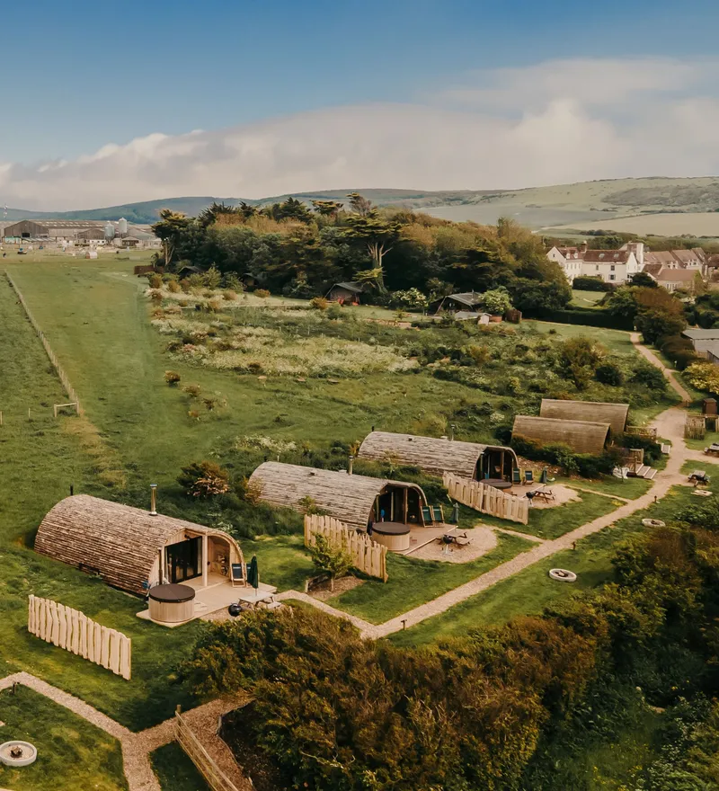 Aerial view of wooden glamping pods beside a green field and countryside village