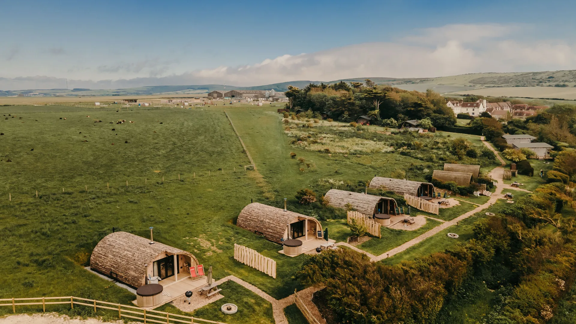 Aerial view of wooden glamping pods beside a green field and countryside village