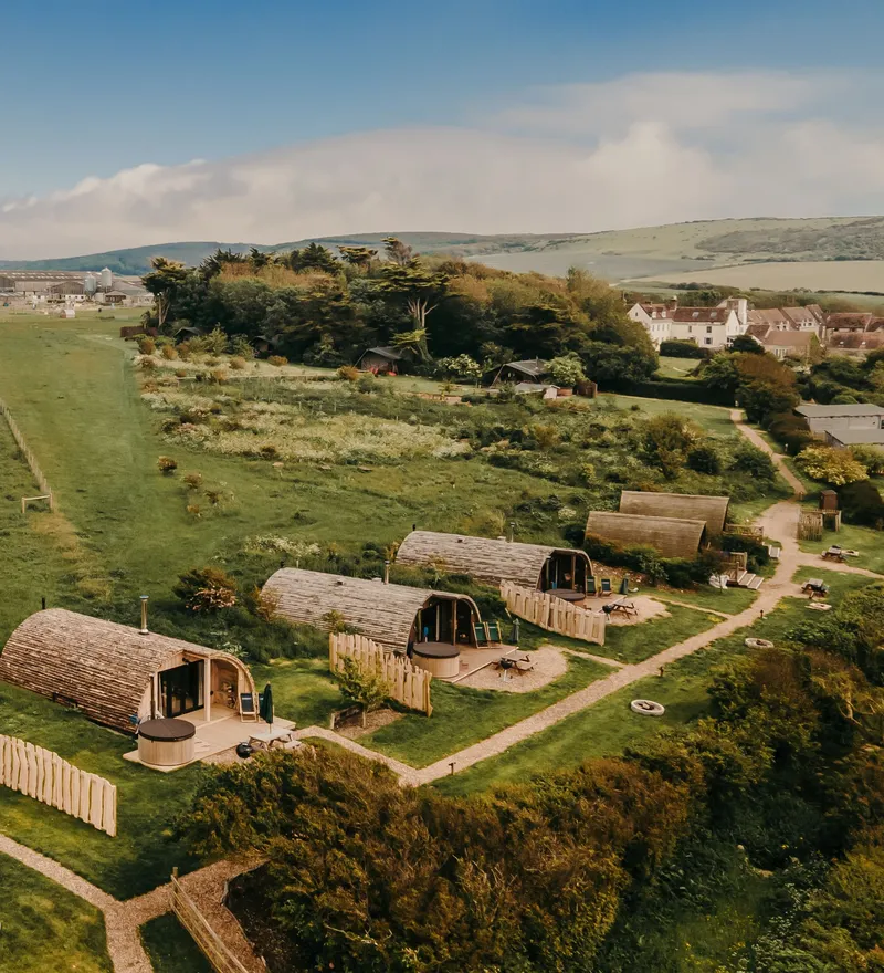 Aerial view of wooden glamping pods beside a green field and countryside village