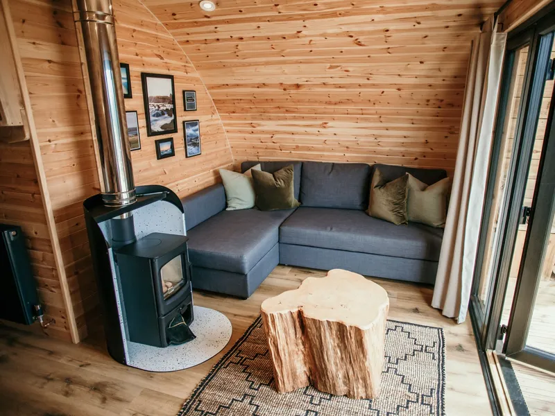 Cosy wooden cabin living area with wood-burning stove, grey corner sofa and log stump coffee table by glass doors