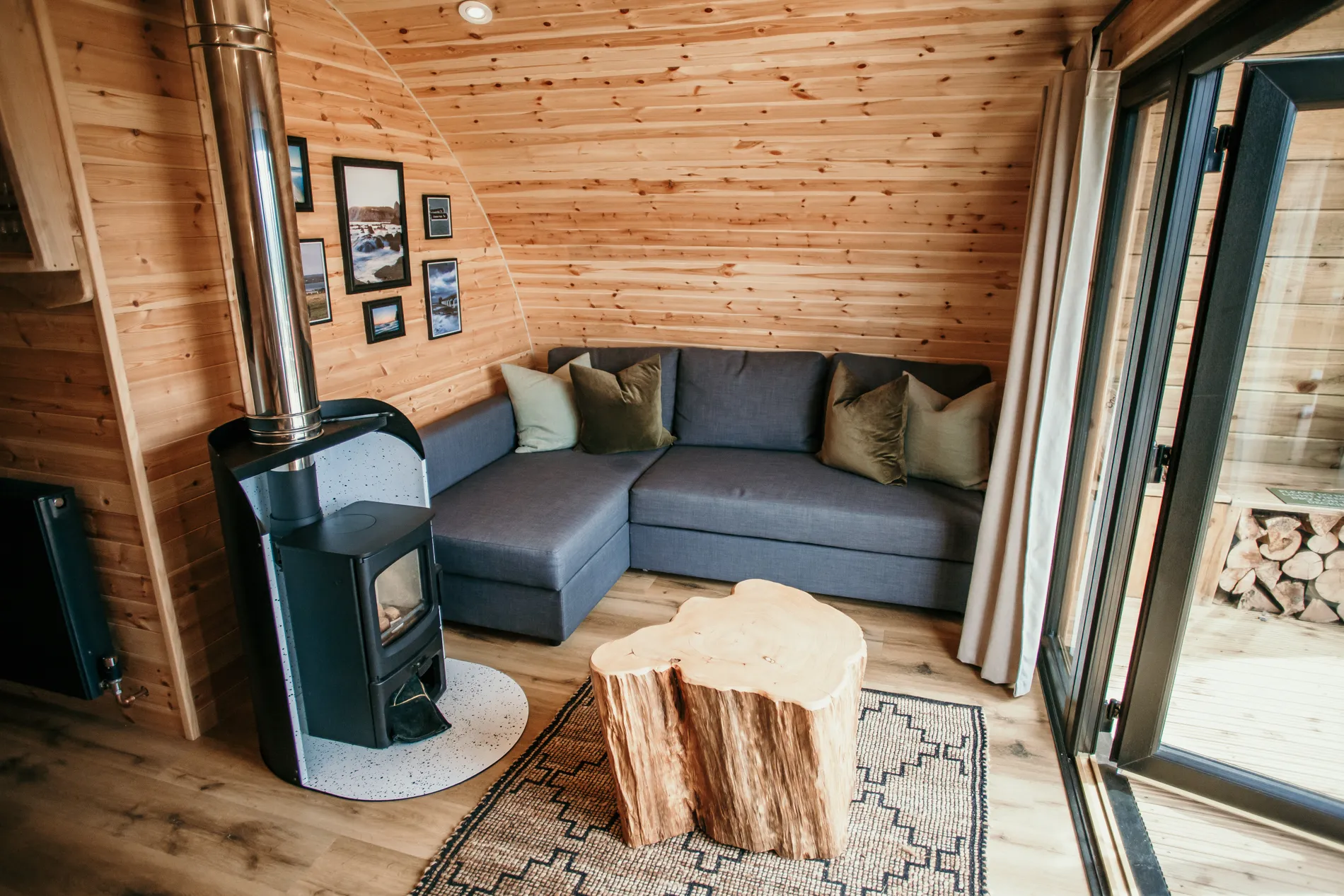 Cosy wooden cabin living area with wood-burning stove, grey corner sofa and log stump coffee table by glass doors