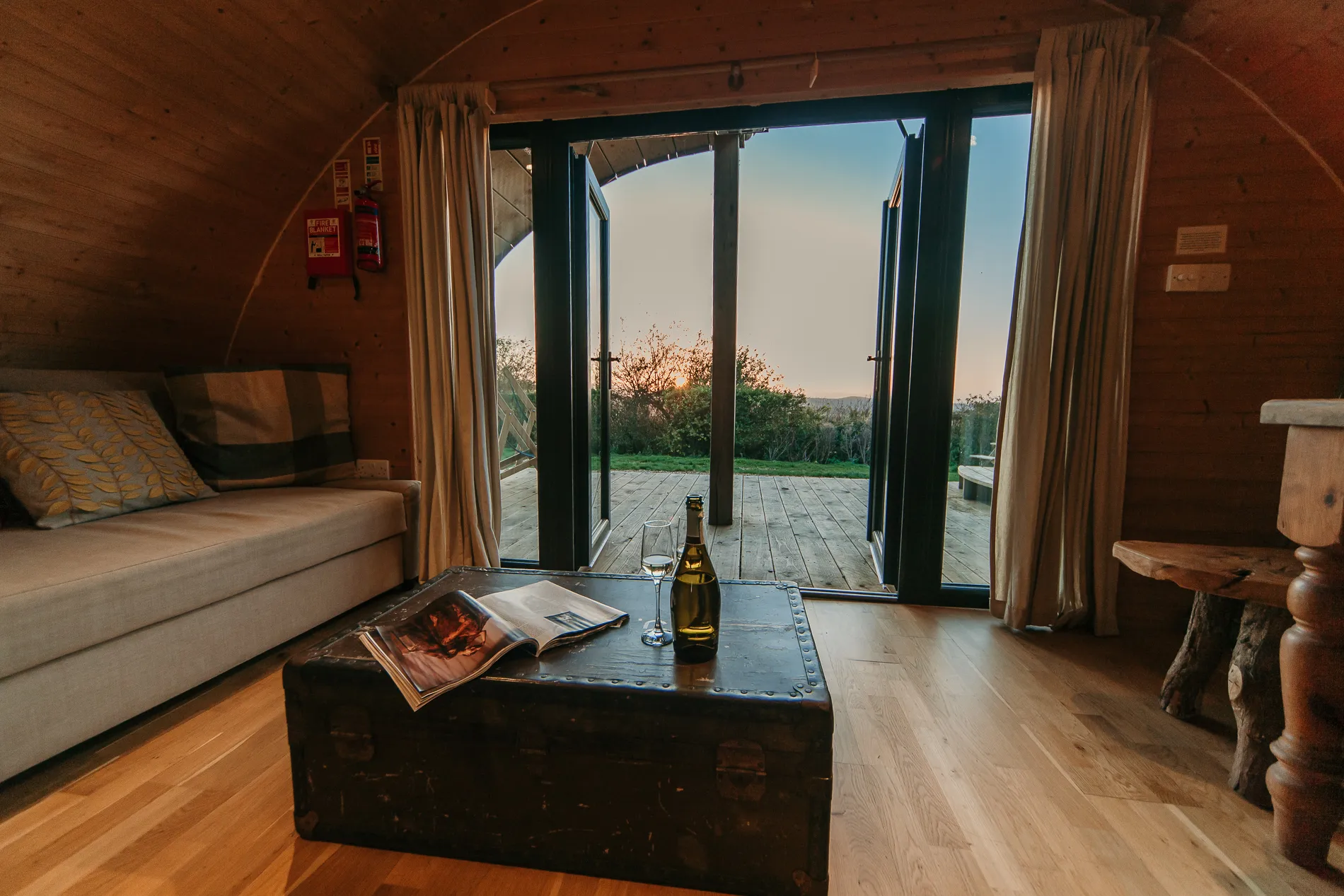 Cosy wooden cabin interior with open double doors to a deck at sunset, with a sofa and a coffee table holding a bottle and glasses