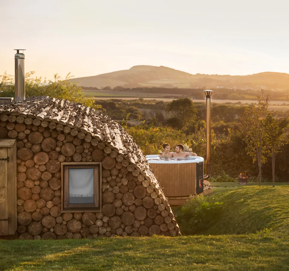 Couple relaxing in a wooden hot tub beside a cosy log cabin at sunset, overlooking rolling countryside