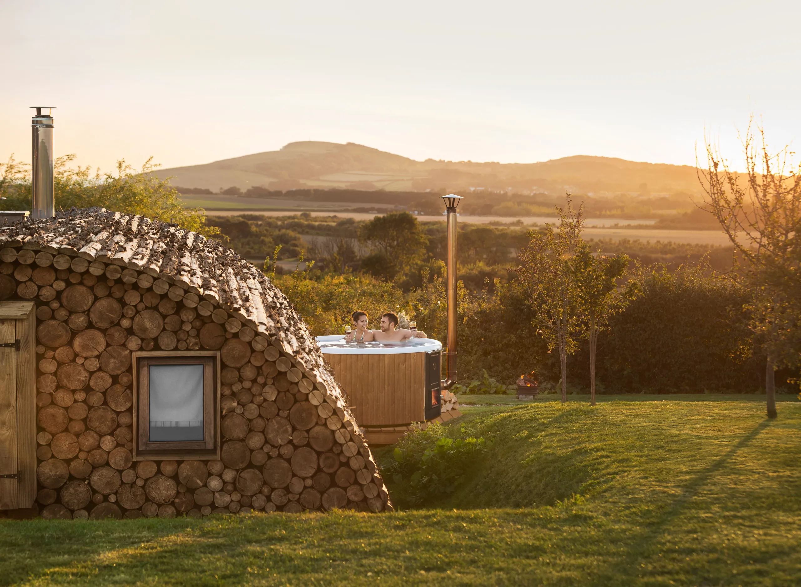 Couple relaxing in a wooden hot tub beside a cosy log cabin at sunset, overlooking rolling countryside