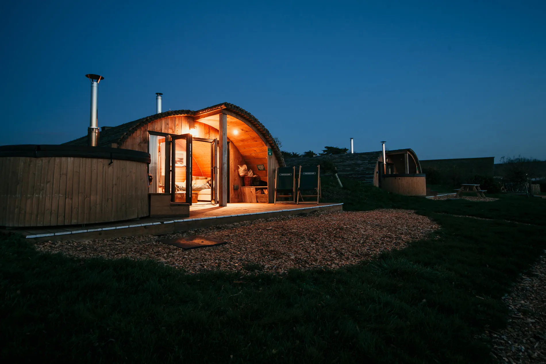 Cosy wooden glamping pod with open doors and lit interior at dusk, beside a hot tub on a grassy campsite
