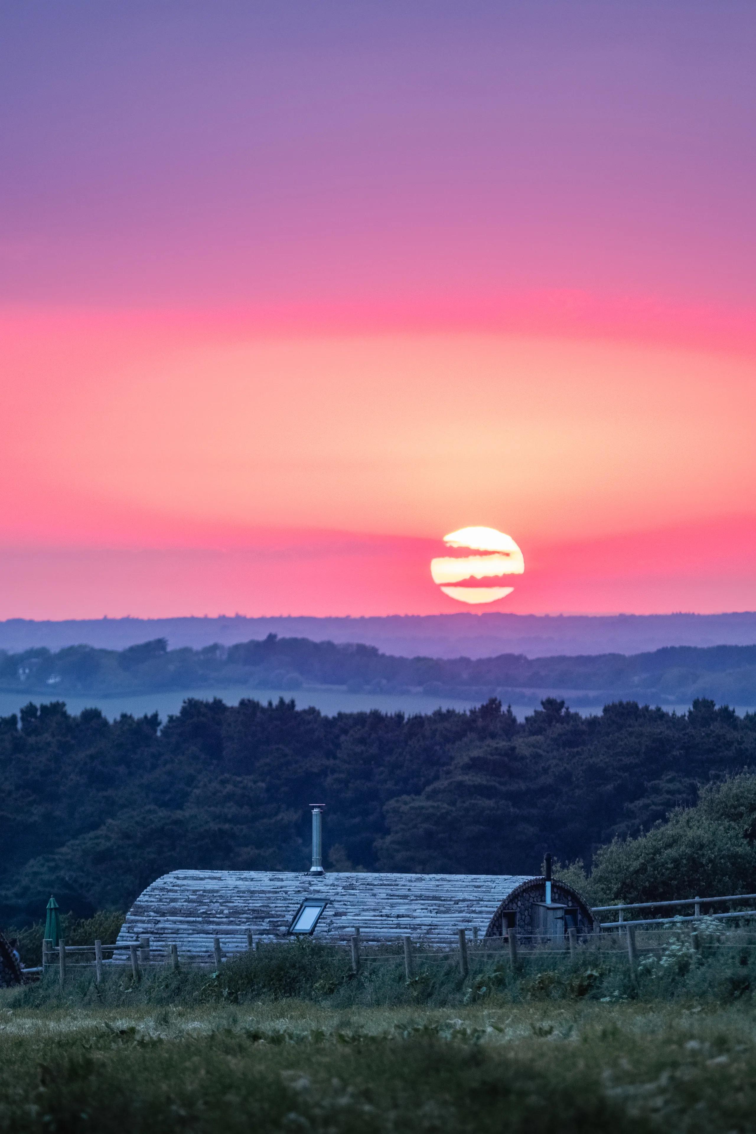 Pink and purple sunset over rolling hills with a cabin roof in the foreground