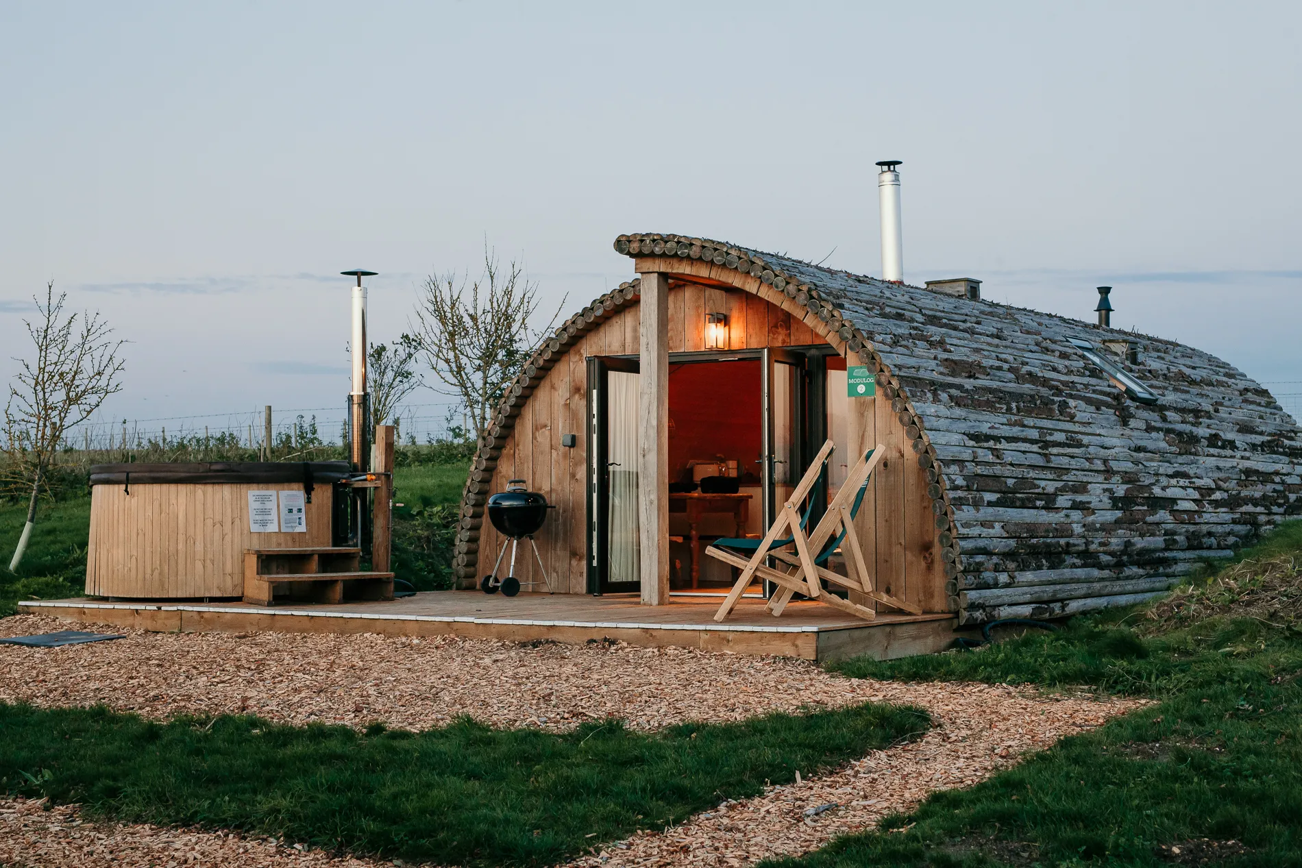 Wooden arch-shaped cabin with open doors, deck chairs, and an outdoor hot tub on a rural campsite at dusk