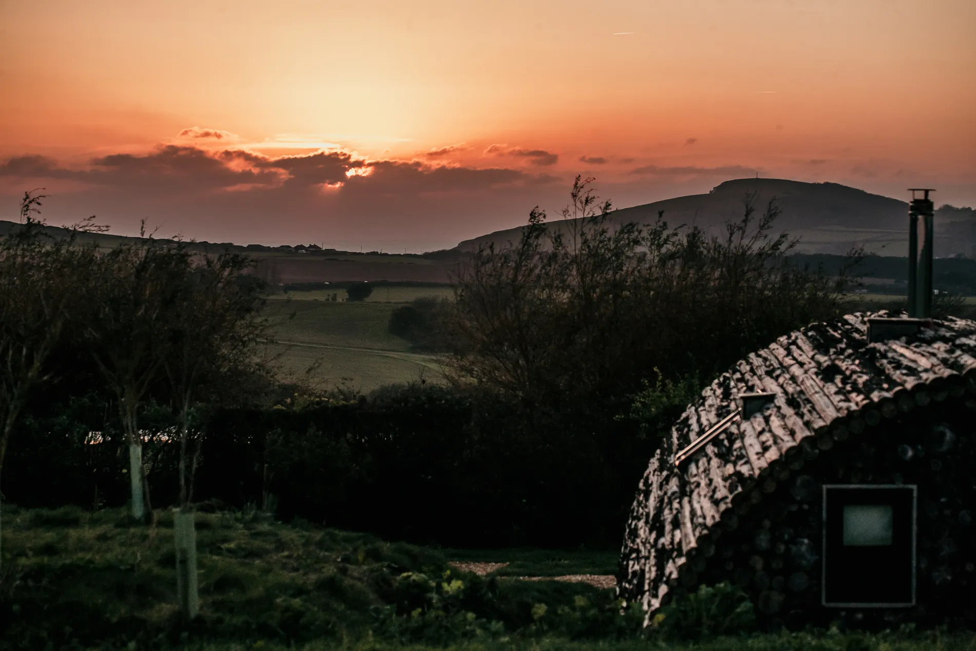 Sunset over rolling countryside with silhouetted hills and a log cabin roof in the foreground