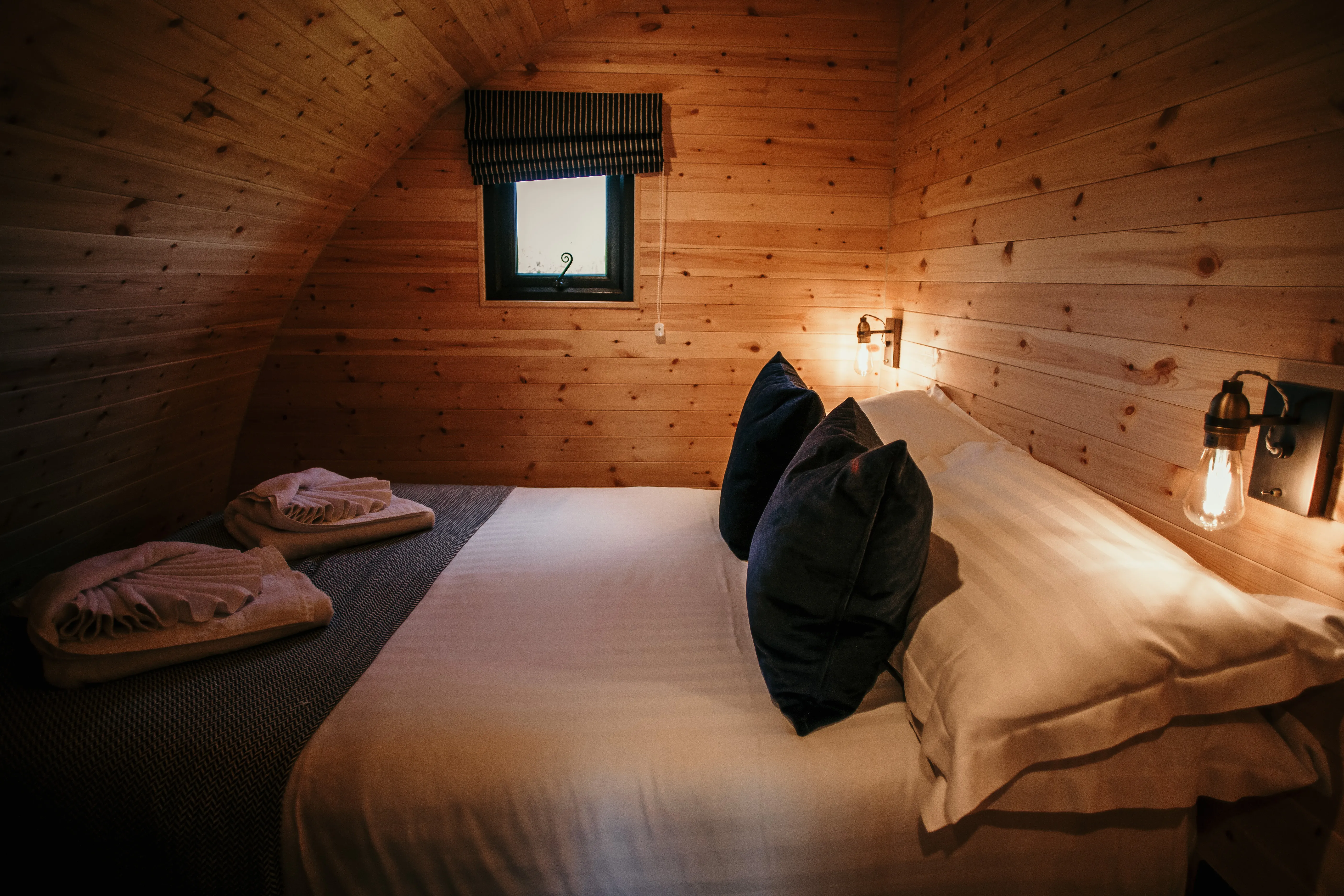 Cosy wooden cabin bedroom with double bed, folded towels, wall lights and small window