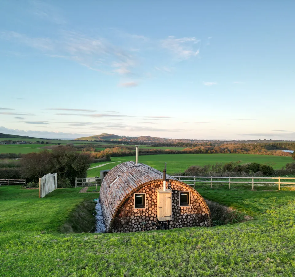 Log-built barrel-shaped cabin in a green field with distant hills under a blue sky