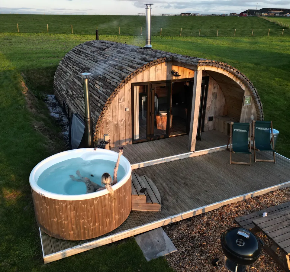 Aerial view of a rustic wooden glamping cabin with a hot tub on a deck in a green countryside setting at sunset