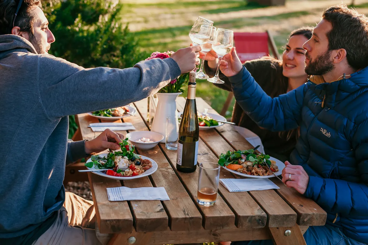 Friends clinking wine glasses over an outdoor picnic table set with dinner plates and a bottle of white wine