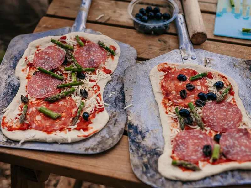 Two uncooked homemade pizzas on metal pizza peels, topped with salami, asparagus, cheese and black olives on a wooden outdoor table
