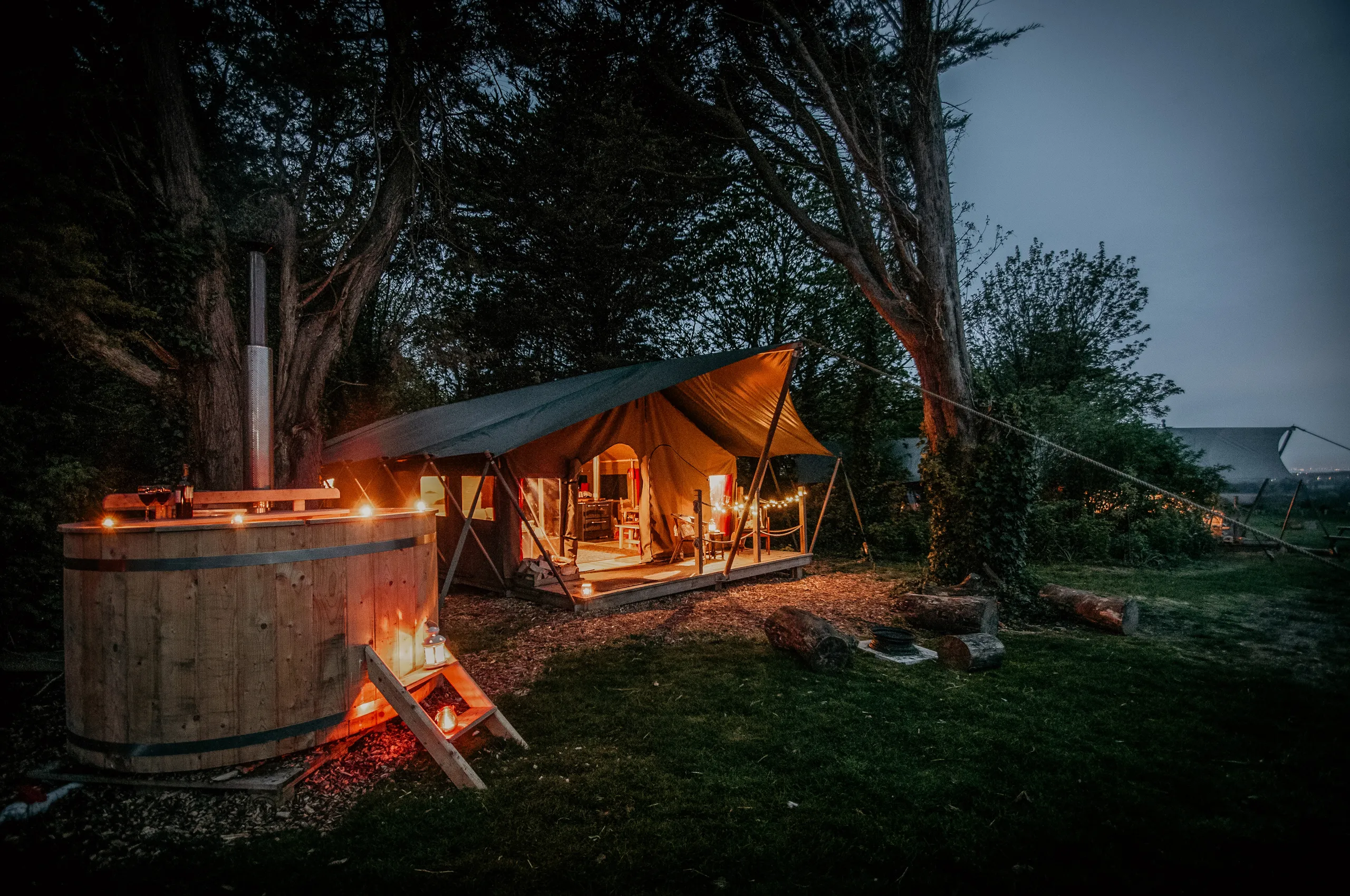Safari-style canvas glamping tent lit with warm lanterns beside a wooden hot tub at dusk