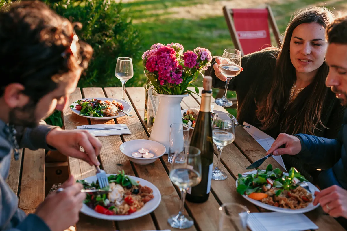 Friends sharing an outdoor meal at a wooden table with wine glasses and a jug of pink flowers