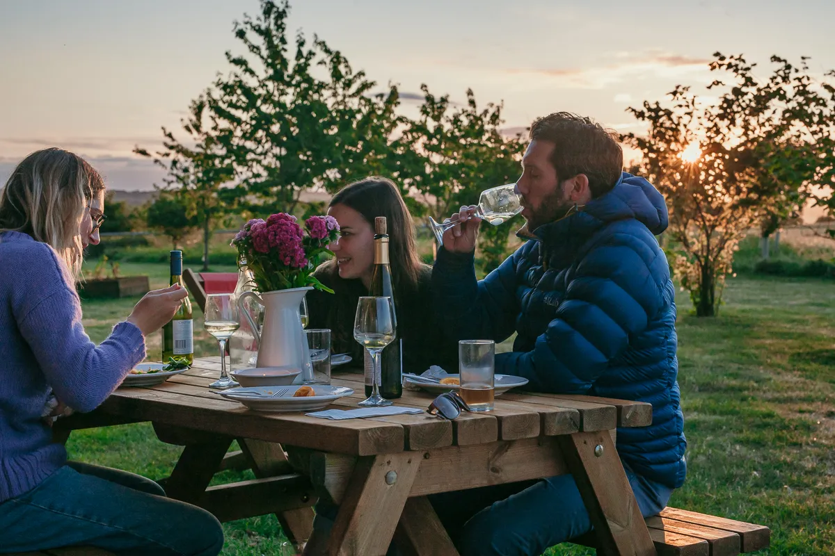 Three people enjoying wine and dinner at a picnic table outdoors at sunset