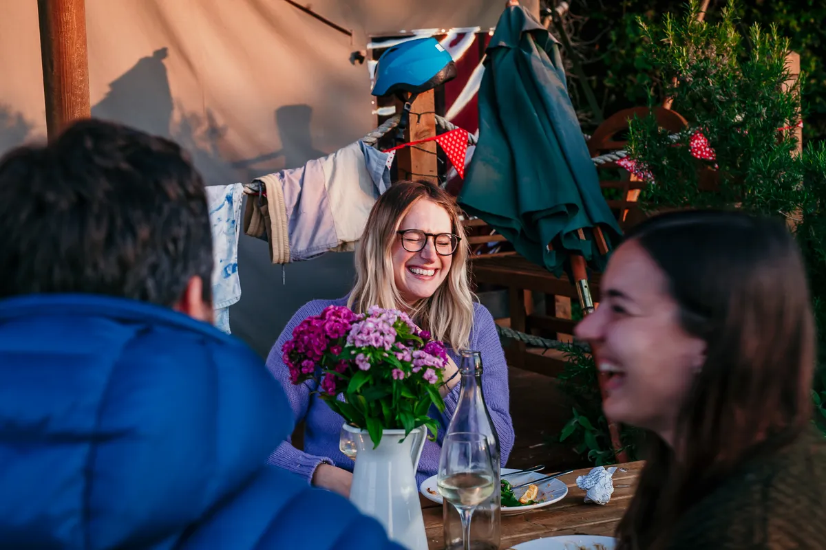 Friends laughing at a wooden table outside a safari tent, with a vase of pink and purple flowers and a bottle of white wine.