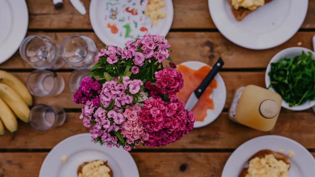 Overhead view of a rustic wooden table set for breakfast with plates of scrambled eggs on toast, smoked salmon, glasses and a bunch of pink flowers in the centre.