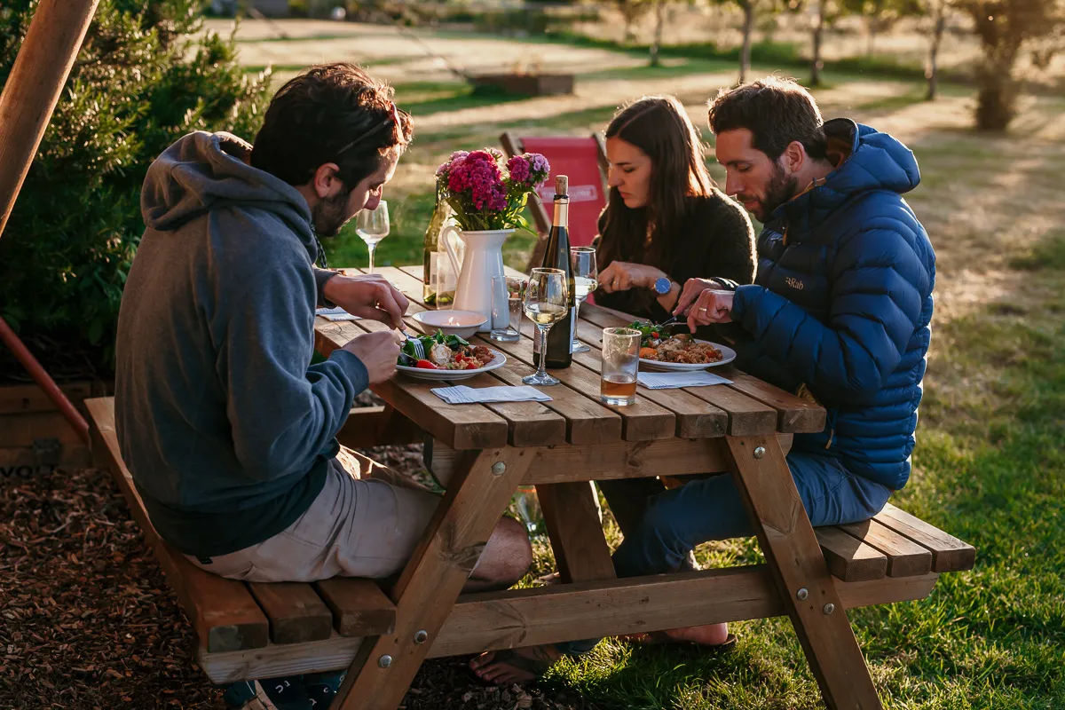 Three friends eating dinner at an outdoor picnic table with wine at sunset