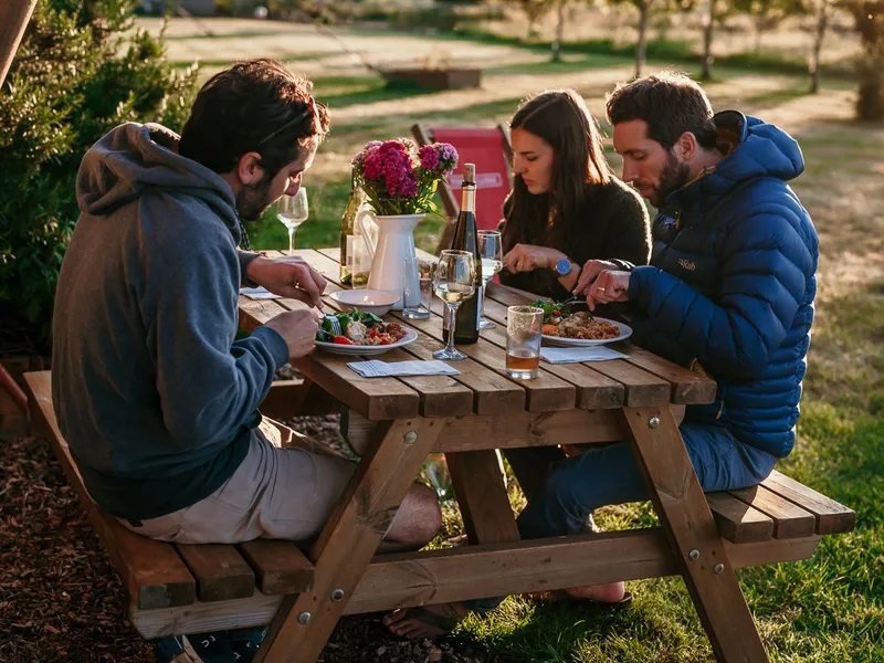 Three friends eating dinner at an outdoor picnic table with wine at sunset