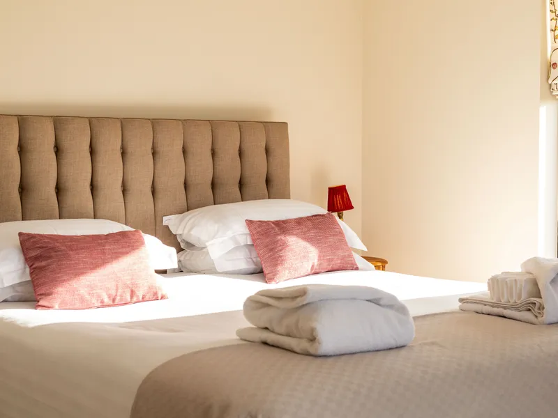 Sunlit bedroom with tufted headboard, white bedding, folded towels and a small red bedside lamp