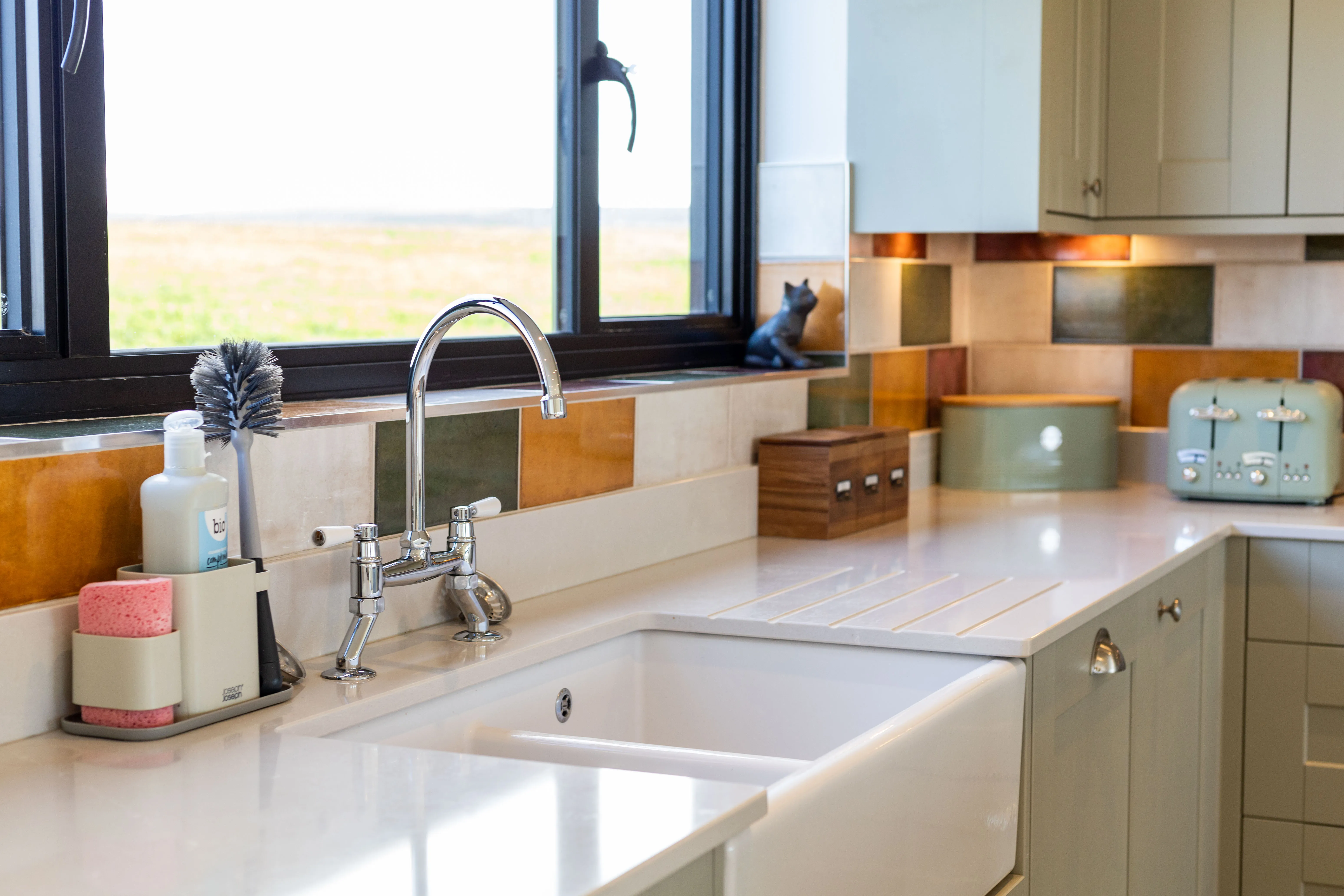 Farmhouse kitchen sink with chrome tap, colourful tiled splashback and pale green units