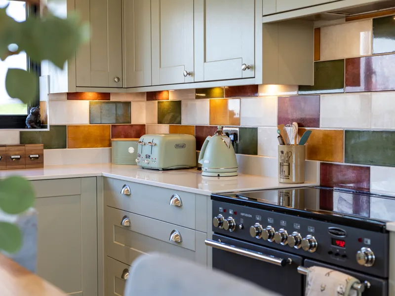 Cosy kitchen corner with sage green cabinets, colourful tiled splashback, and matching toaster and kettle on a white worktop beside a range cooker.