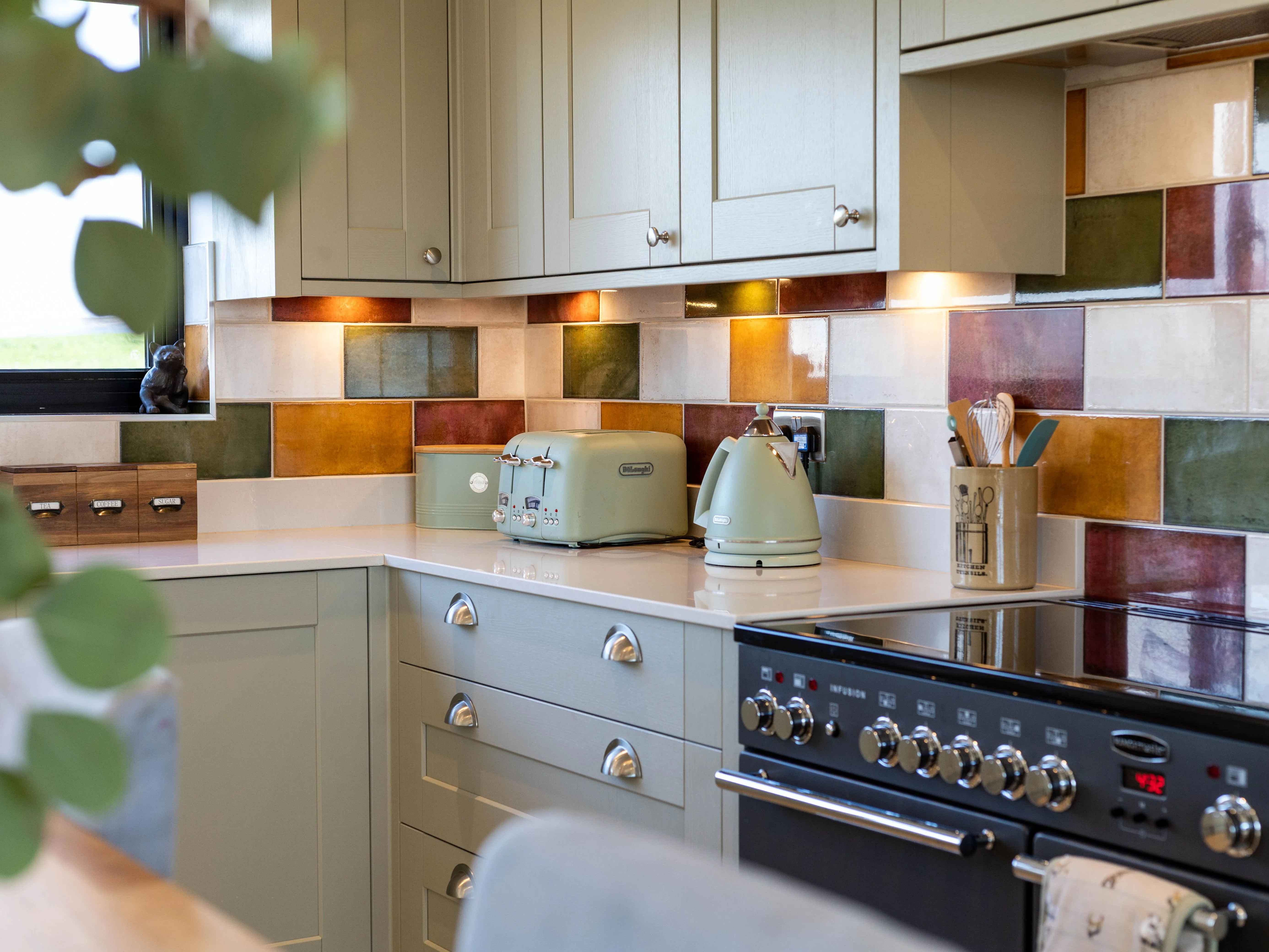 Cosy kitchen corner with sage green cabinets, colourful tiled splashback, and matching toaster and kettle on a white worktop beside a range cooker.