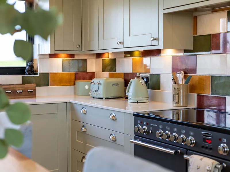 Cosy kitchen corner with sage green cabinets, colourful tiled splashback, and matching toaster and kettle on a white worktop beside a range cooker.