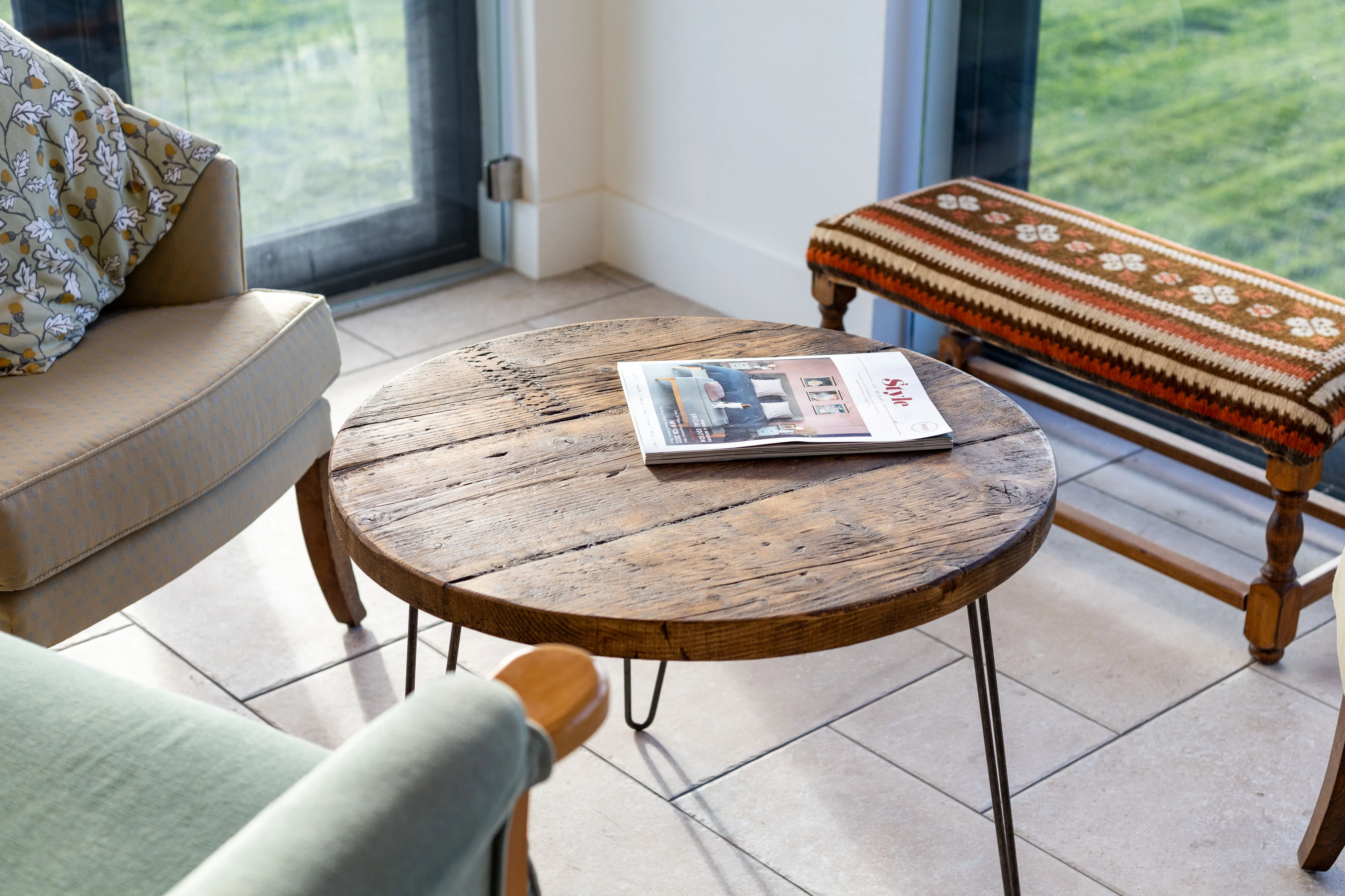 Rustic round wooden coffee table with a magazine, beside an armchair and patterned bench by large windows