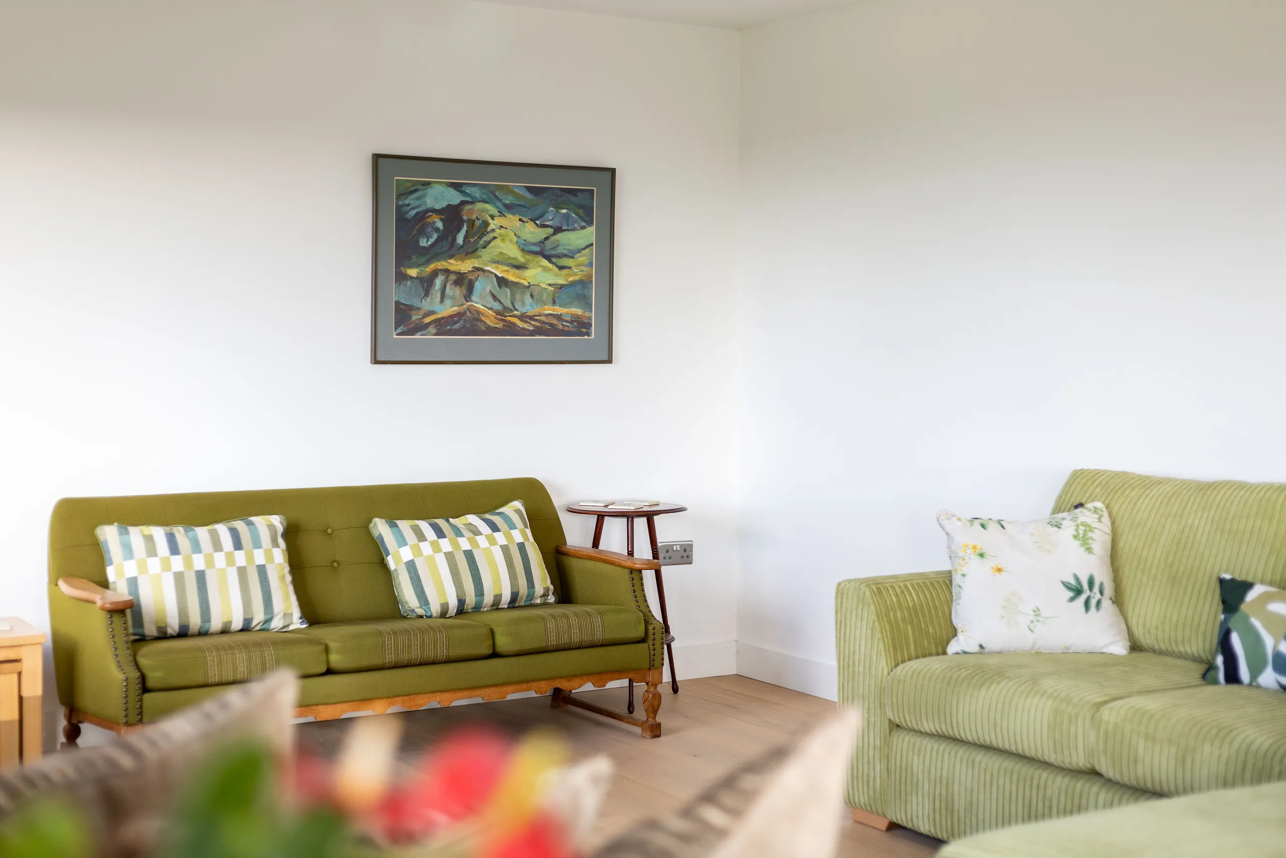 Living room with green sofa and armchair, patterned cushions and a framed landscape painting on a white wall