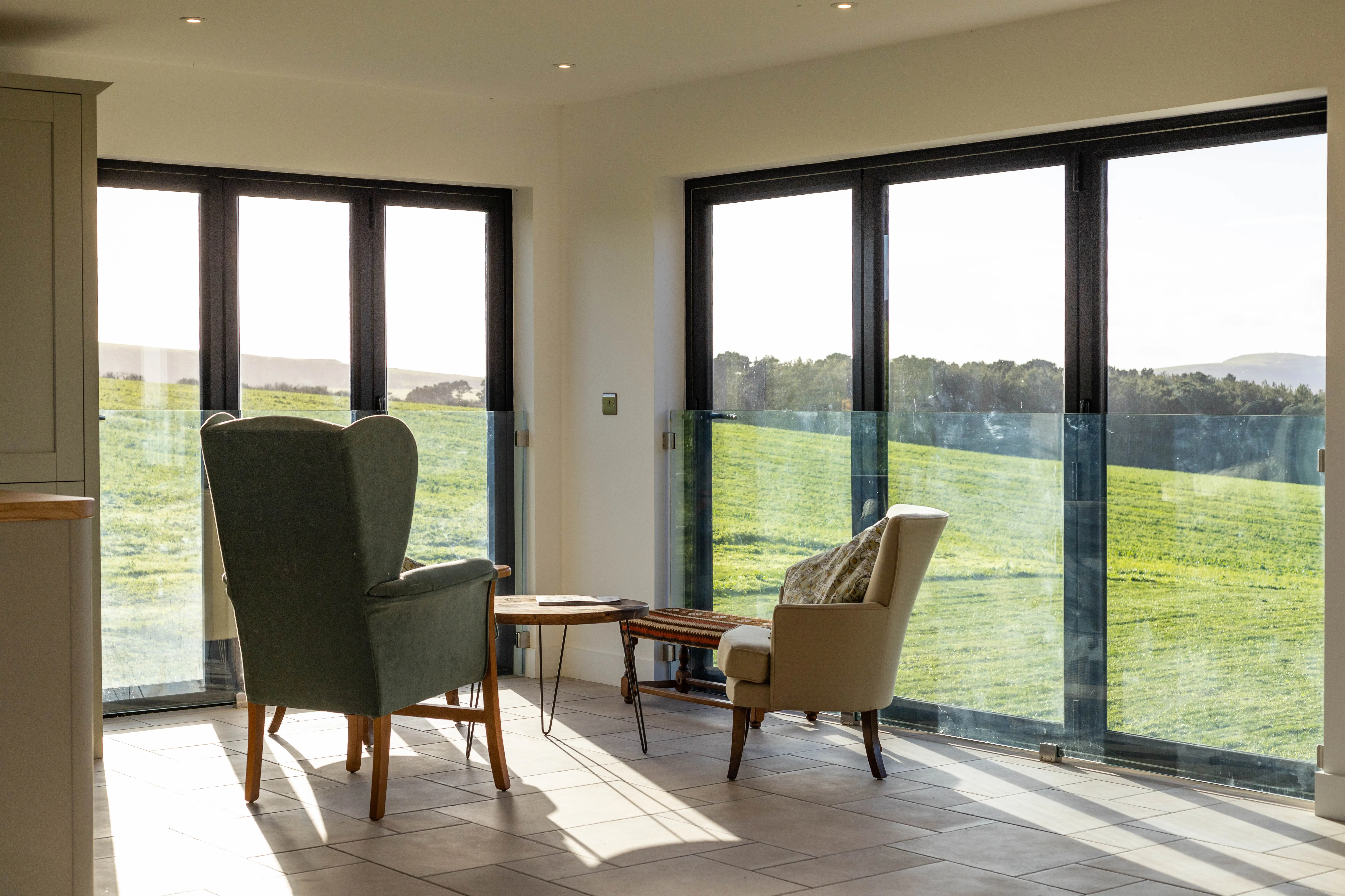 Sunlit seating area with armchairs beside floor-to-ceiling windows overlooking green countryside