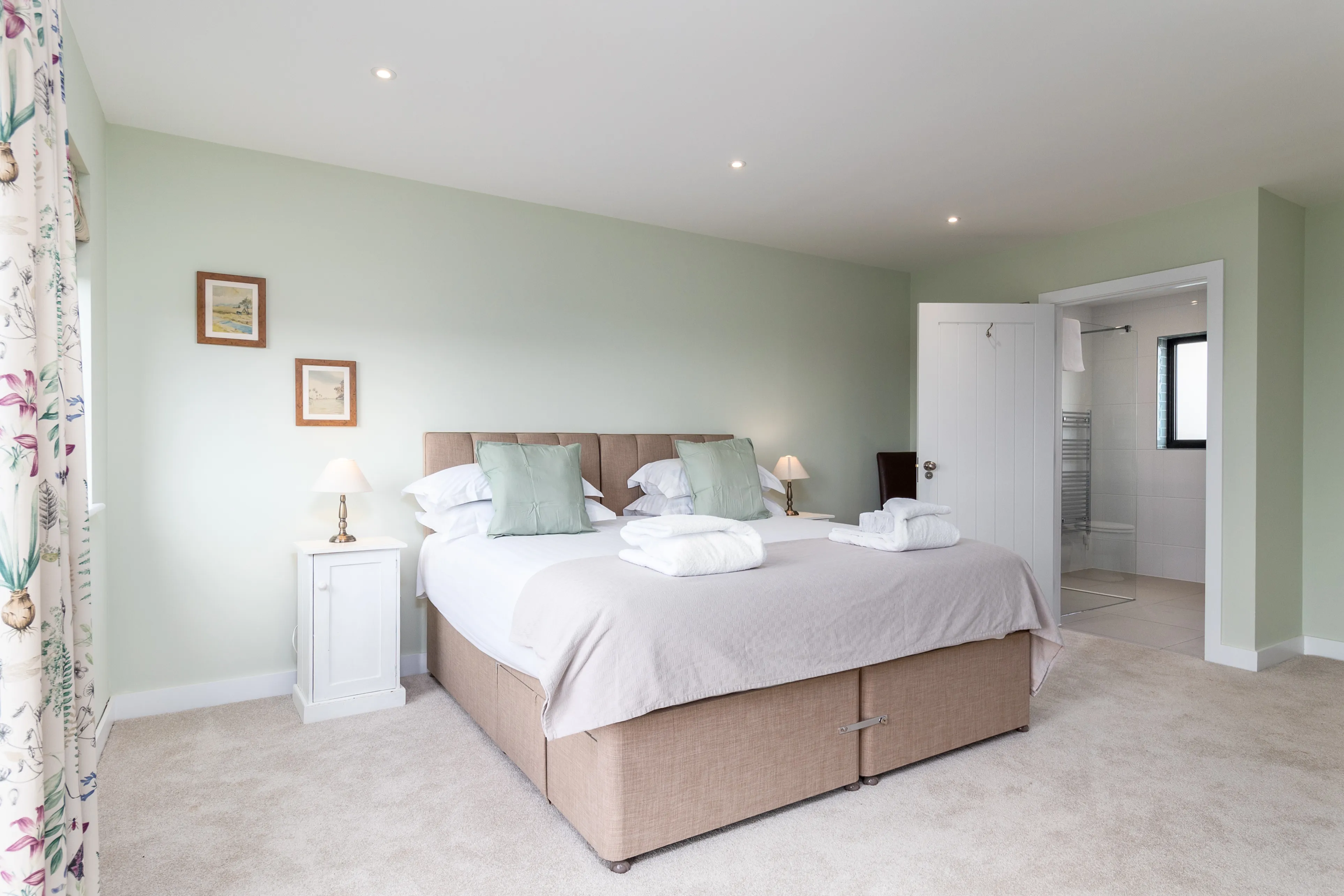 Bright double bedroom with pale green walls, upholstered bed, bedside lamps and open en-suite bathroom door