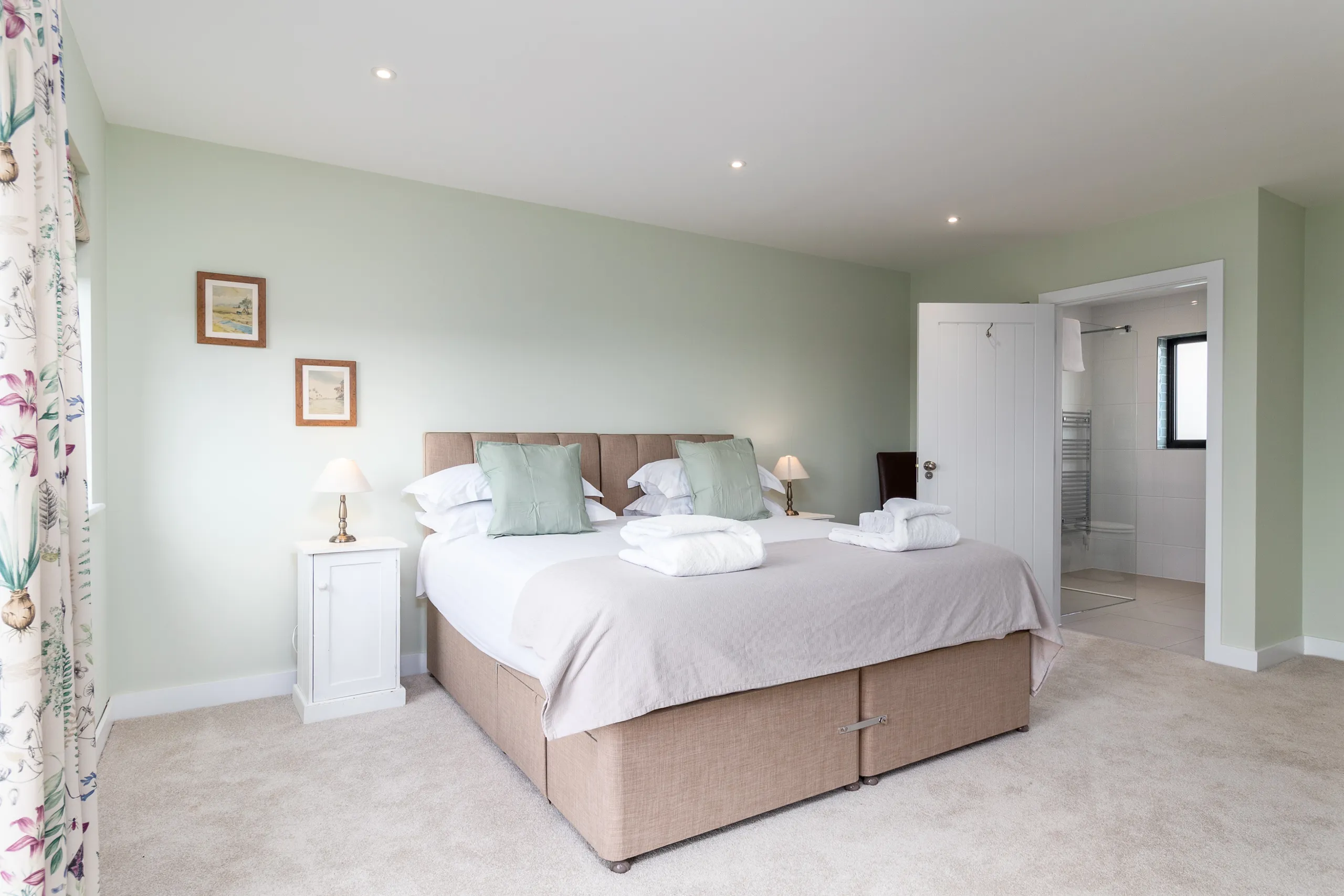 Bright double bedroom with pale green walls, upholstered bed, bedside lamps and open en-suite bathroom door
